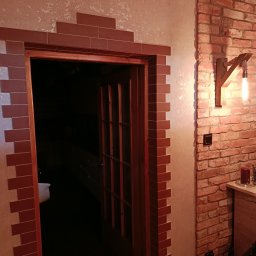 Otwarta drewniana stolarka drzwiowa z dekoracyjnym obramowaniem z cegieł, przylegająca do ściany z cegły z drewnianą lampą. Widoczny fragment wnętrza.