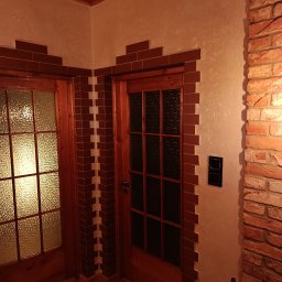 MIRBUD Mirosław Pastuszek - Wykończenie wnętrza z drewnianymi drzwiami, obramowanymi dekoracyjną cegłą. Ściany pokryte jasnym tynkiem strukturalnym, drewniana podłoga i sufit. Widoczny fragment ściany z cegły.