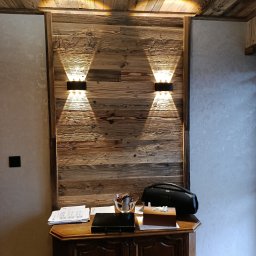 MIRBUD Mirosław Pastuszek - Drewniana ściana z dwoma symetrycznie umieszczonymi kinkietami, oświetlającymi przestrzeń nad komodą z akcesoriami. Ciepłe światło podkreśla fakturę drewna.