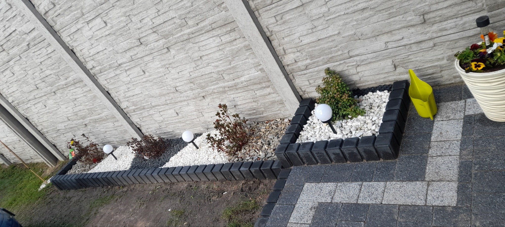 Ogródek z obrzeżami z kostki, białym żwirem, roślinami i lampami kulami, przylegający do szarego, panelowego ogrodzenia. Donica z kwiatami i konewka w tle.