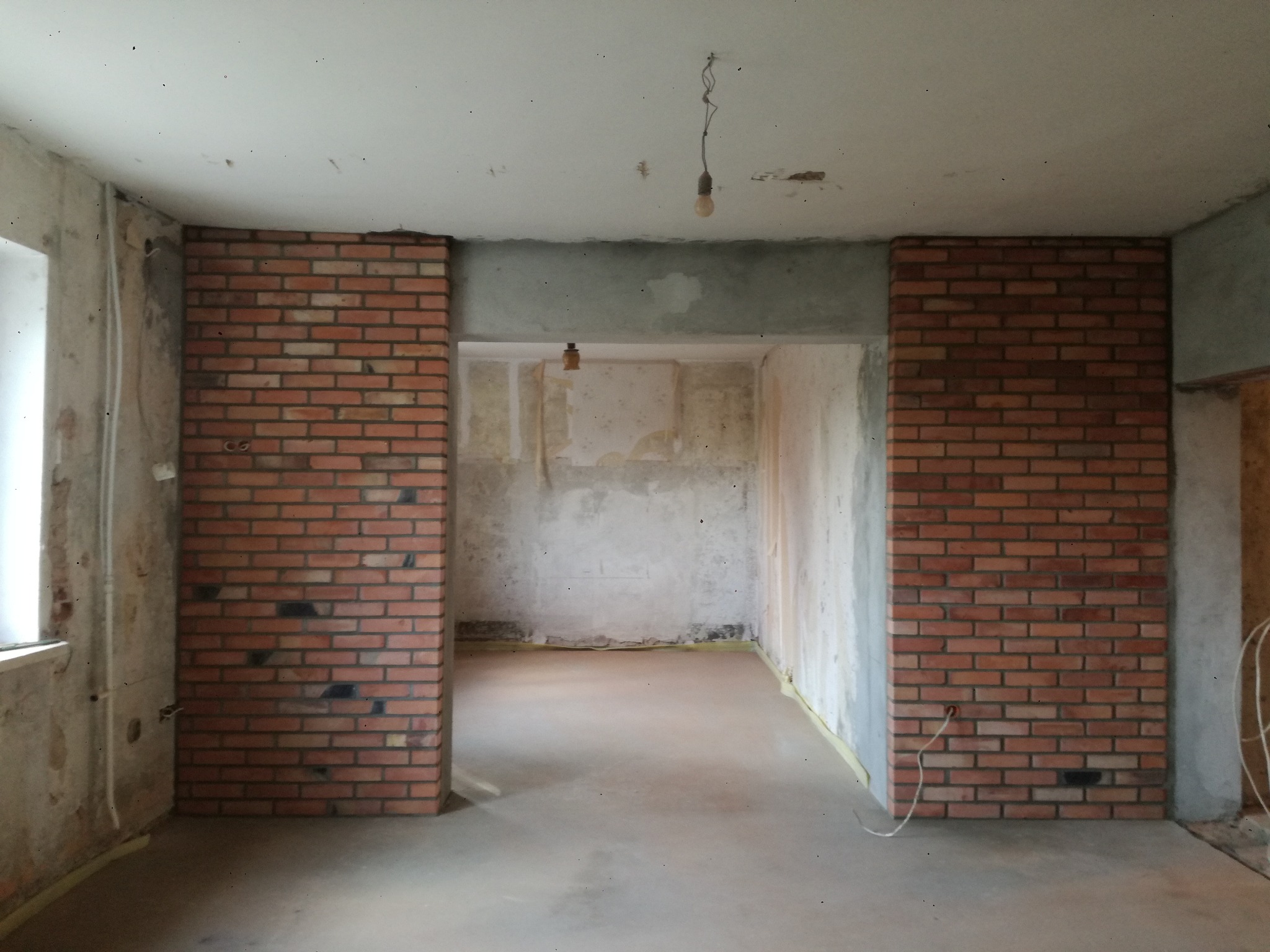 Wnętrze w trakcie wykończenia: fragment ściany z cegły klinkierowej, surowy beton, widoczne instalacje. Oświetlenie tymczasowe, podłoga wylewka.