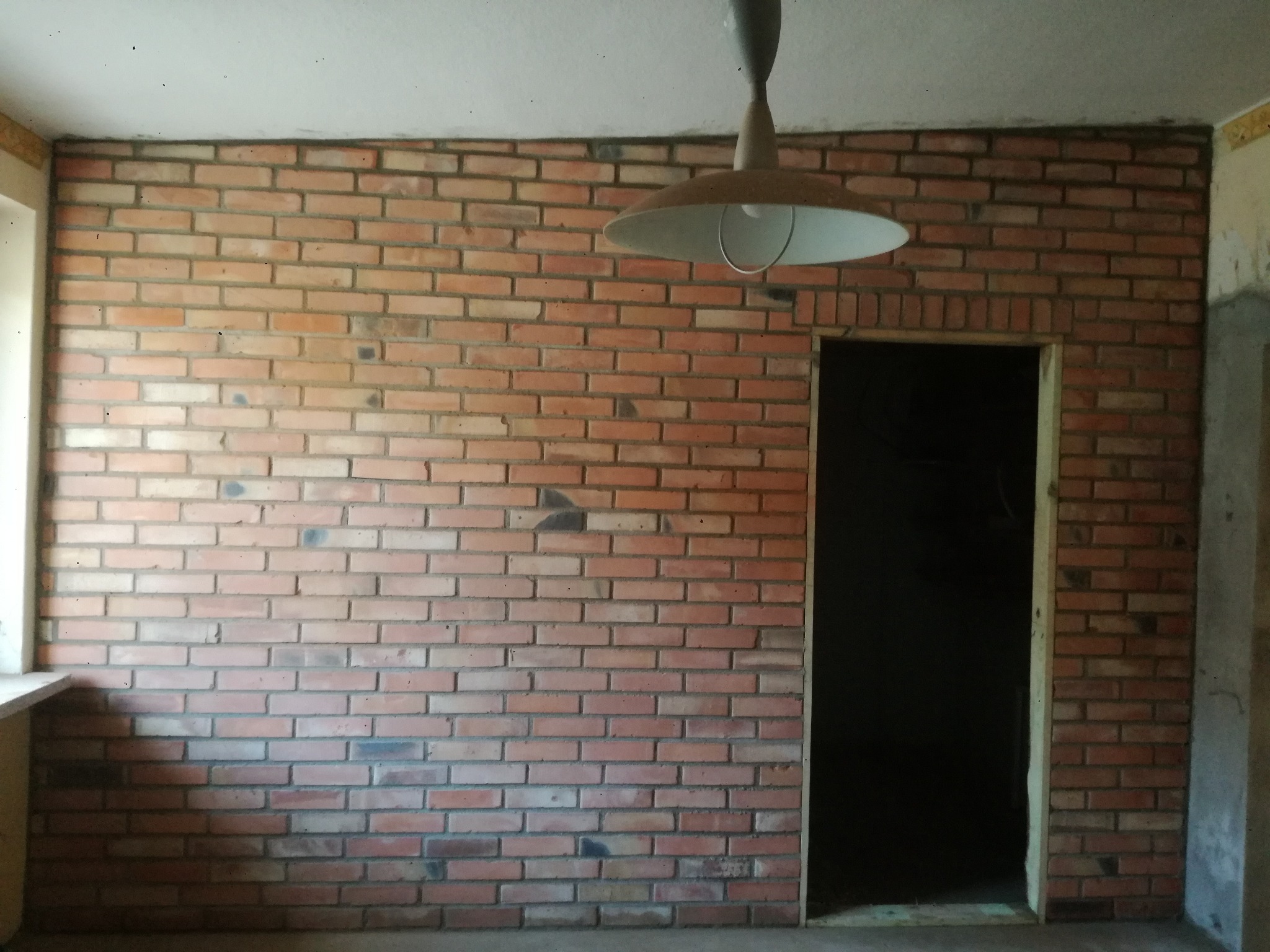 Ściana z cegły klinkierowej w pomieszczeniu z widocznym oknem i otworem drzwiowym, oświetlona wiszącą lampą. Wykończenie wnętrza w stylu industrialnym.
