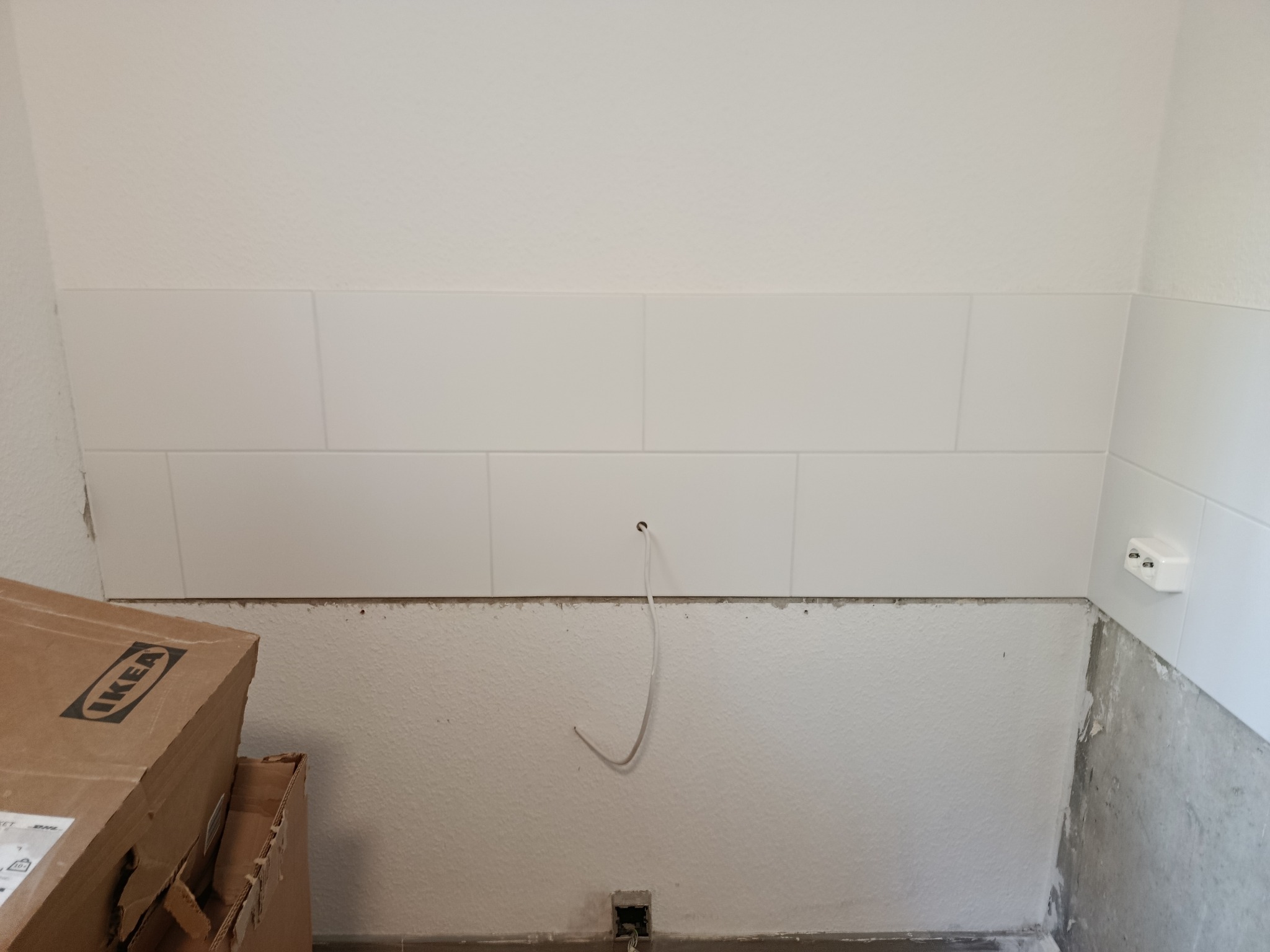 Białe, prostokątne płytki ułożone na ścianie w łazience, widoczne przewody elektryczne i karton IKEA, sugerujące etap prac wykończeniowych.