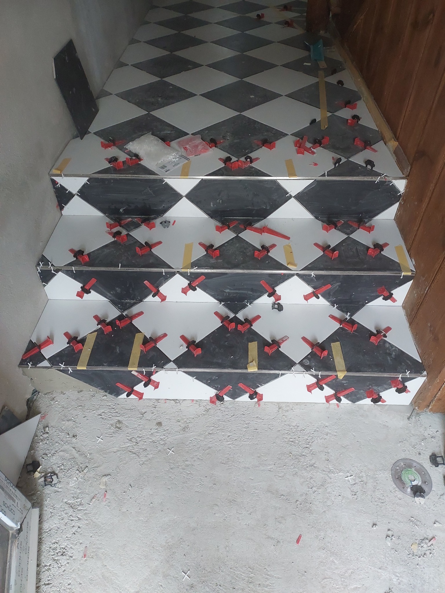 Układanie czarno-białych płytek w romby na schodach z użyciem czerwonych klipsów poziomujących. Widoczne fragmenty płytek i narzędzia.