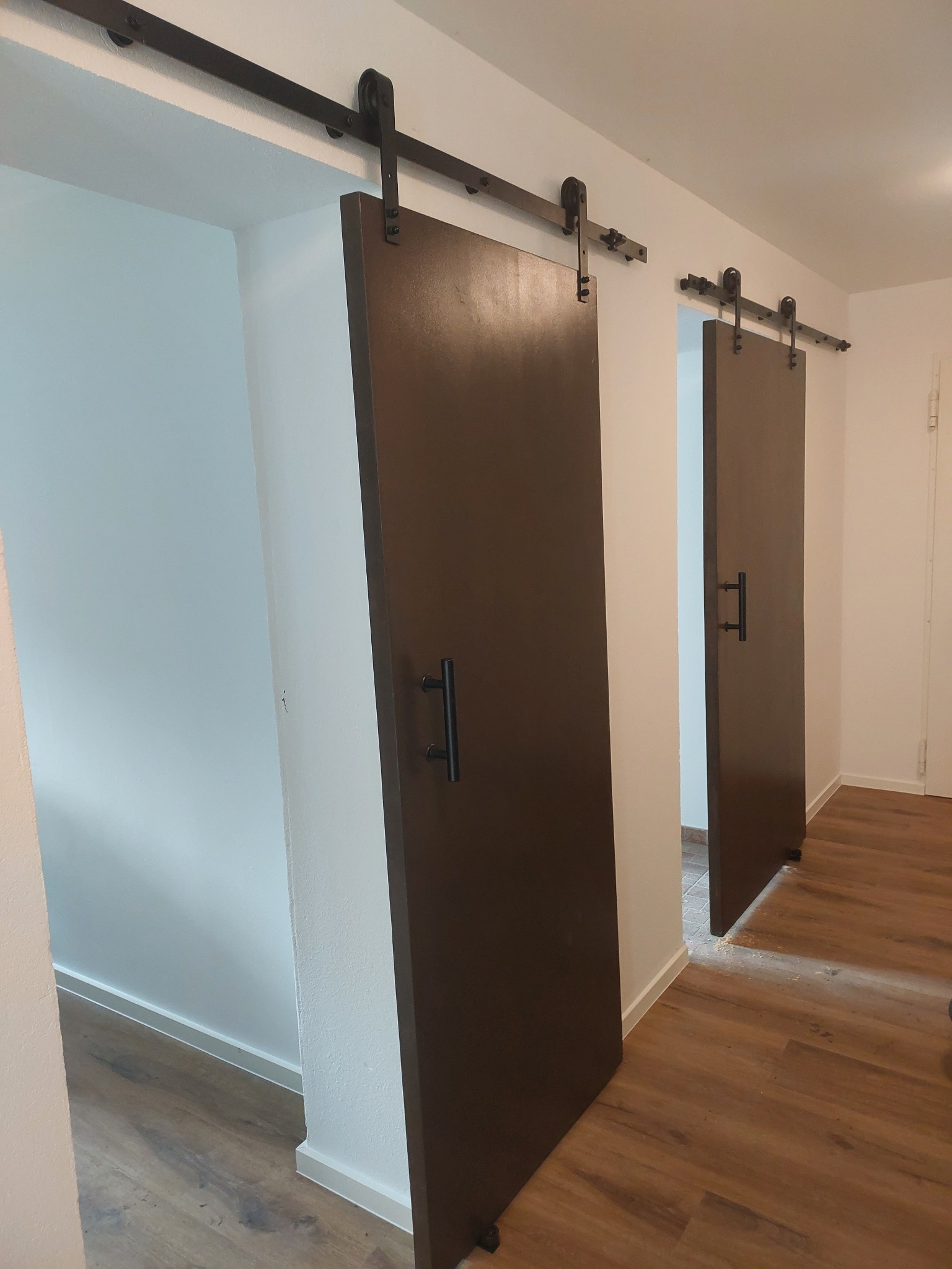 Dwa brązowe, przesuwne drzwi stodołowe z czarnymi uchwytami i szynami, zamontowane w białej ścianie. Widoczna podłoga z paneli i fragment drugiego pomieszczenia.