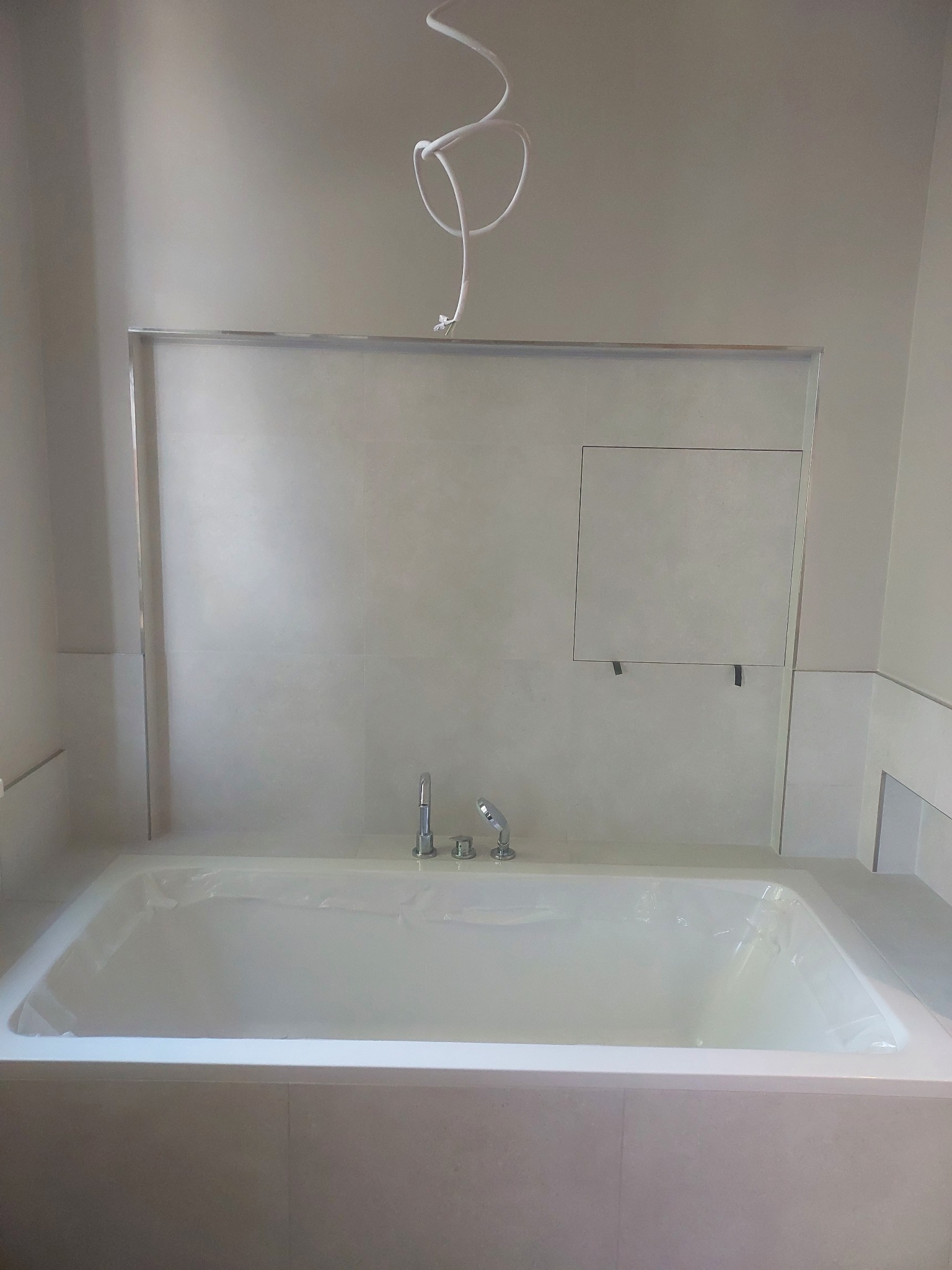 Wykończona łazienka z wanną w zabudowie, jasne płytki, bateria wannowa. Widoczne przewody elektryczne. Klapka rewizyjna na ścianie.