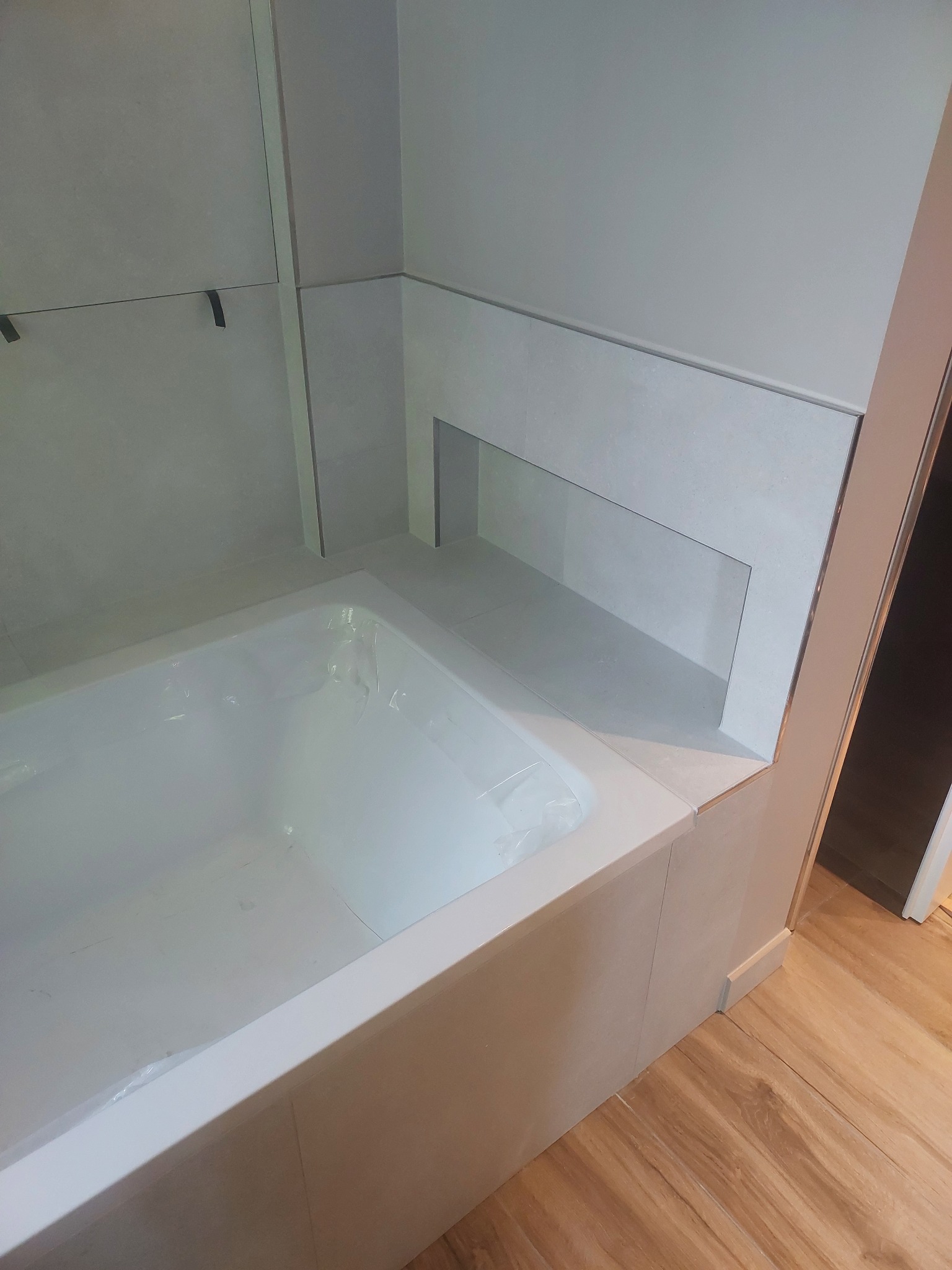 Nowoczesna, wykafelkowana wanna z wnęką na kosmetyki, zabezpieczona folią ochronną. Jasne płytki i drewnopodobna podłoga tworzą minimalistyczny, czysty design łazienki.