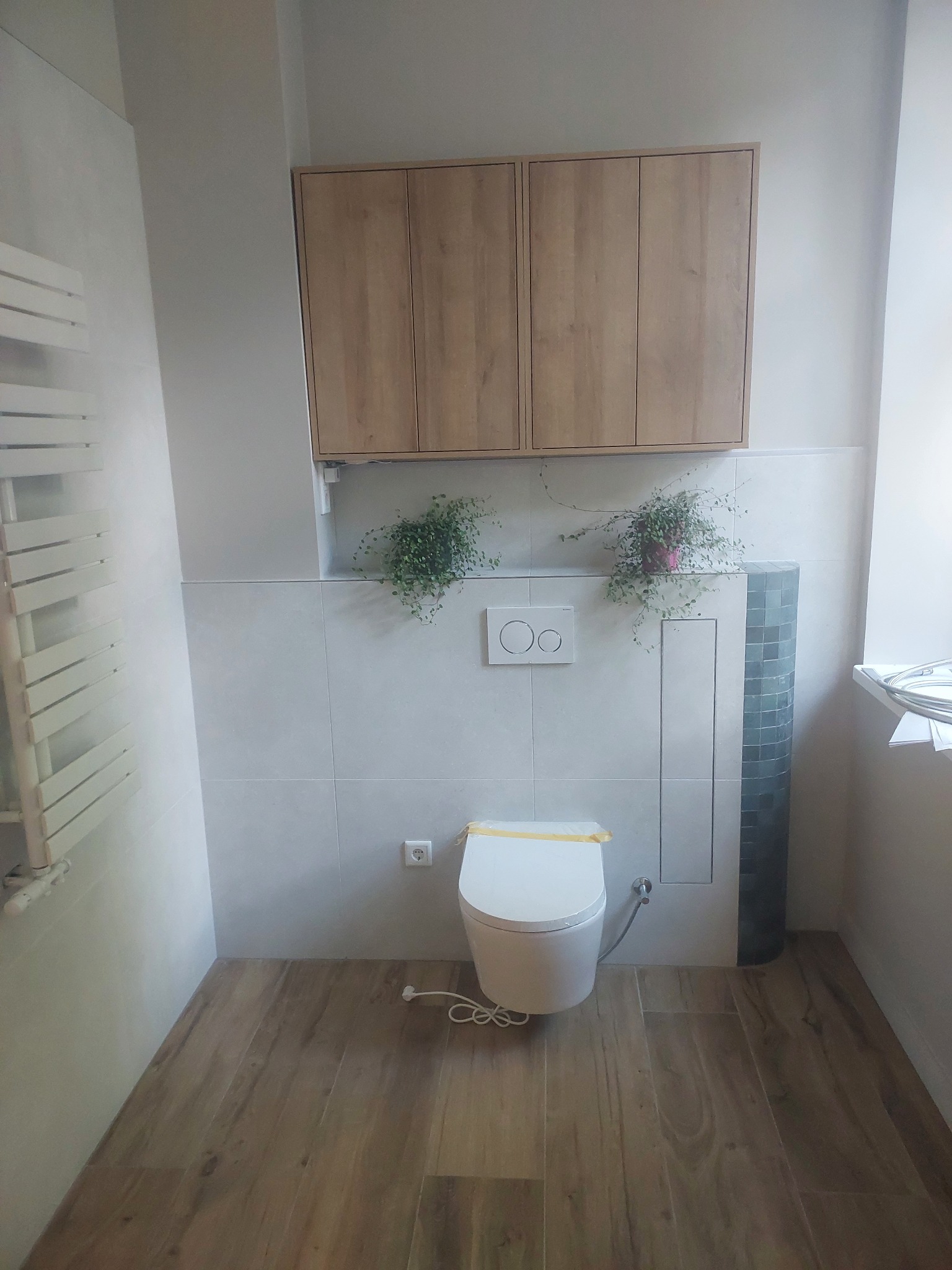 Wykończona łazienka z podwieszaną toaletą, drewnianą szafką nad nią i drewnopodobną podłogą. Na ścianie płytki imitujące kamień i dekoracyjne rośliny.