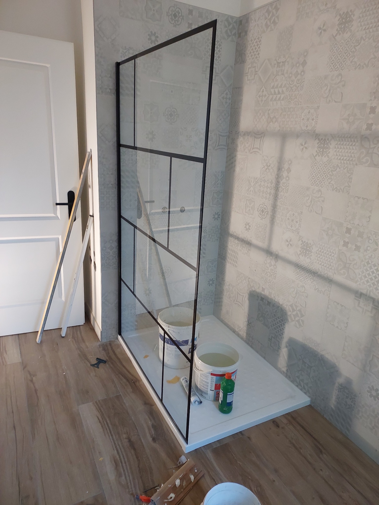 Nowoczesna kabina prysznicowa z czarnymi profilami na tle wzorzystych płytek. Widoczne narzędzia i akcesoria montażowe, sugerujące etap instalacji w łazience.