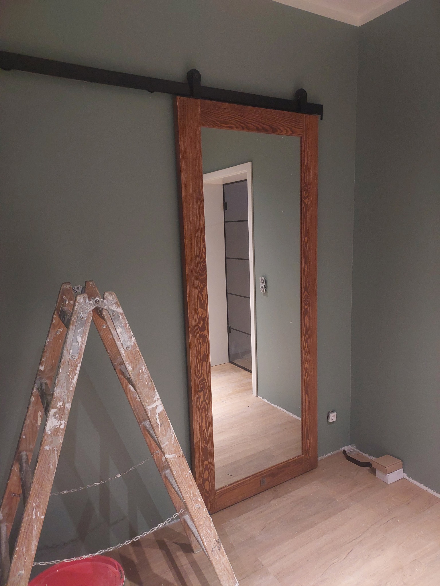 Drewniane drzwi przesuwne z lustrem, zamontowane na szynie, stojące na podłodze z paneli. Obok drabina malarska i ściana w odcieniach szarości. Widoczny fragment wnętrza.