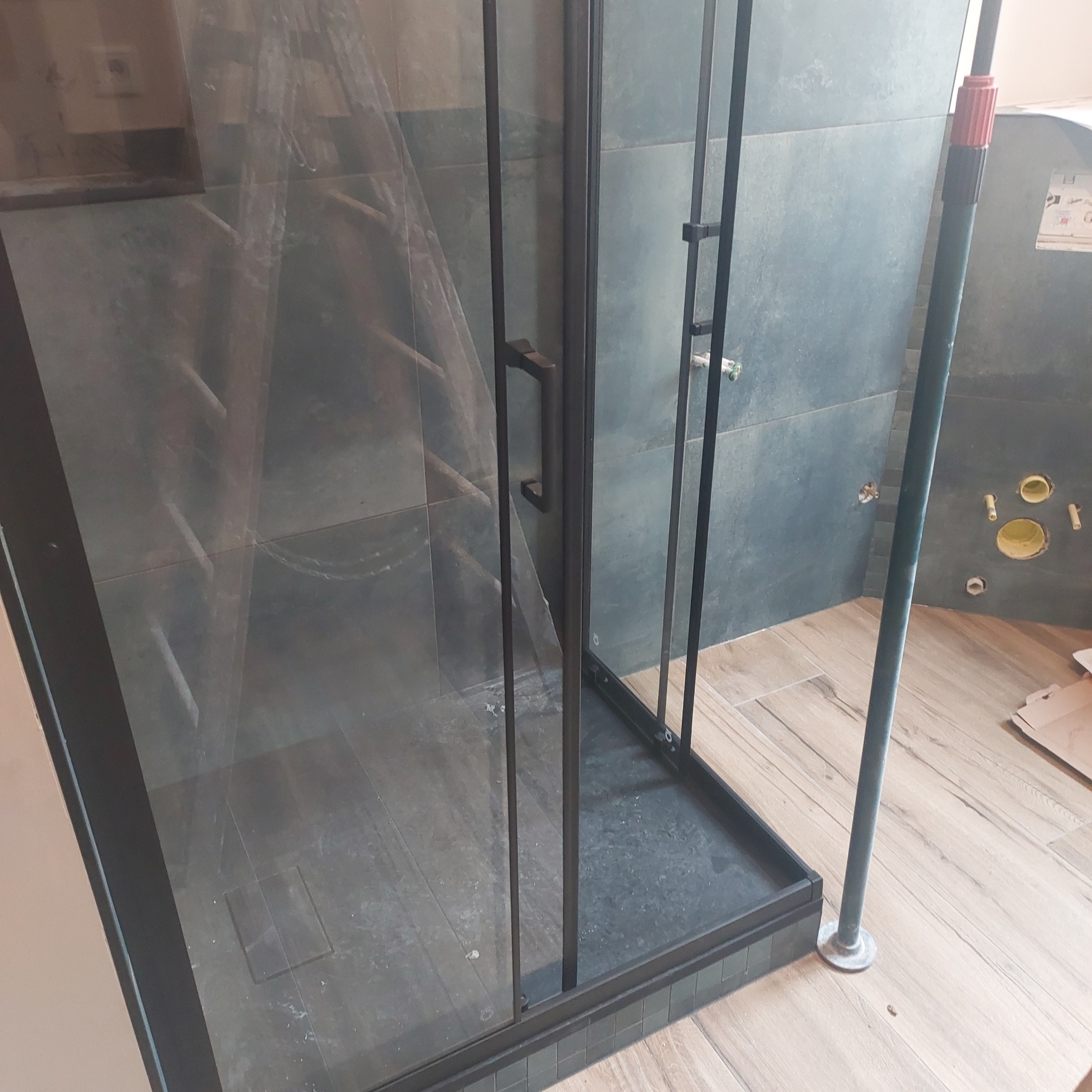 Kabina prysznicowa z czarnymi profilami na tle szarych płytek. Widoczne przyłącza wodne i fragment podłogi imitującej drewno. Częściowo zakryta folią ochronną.