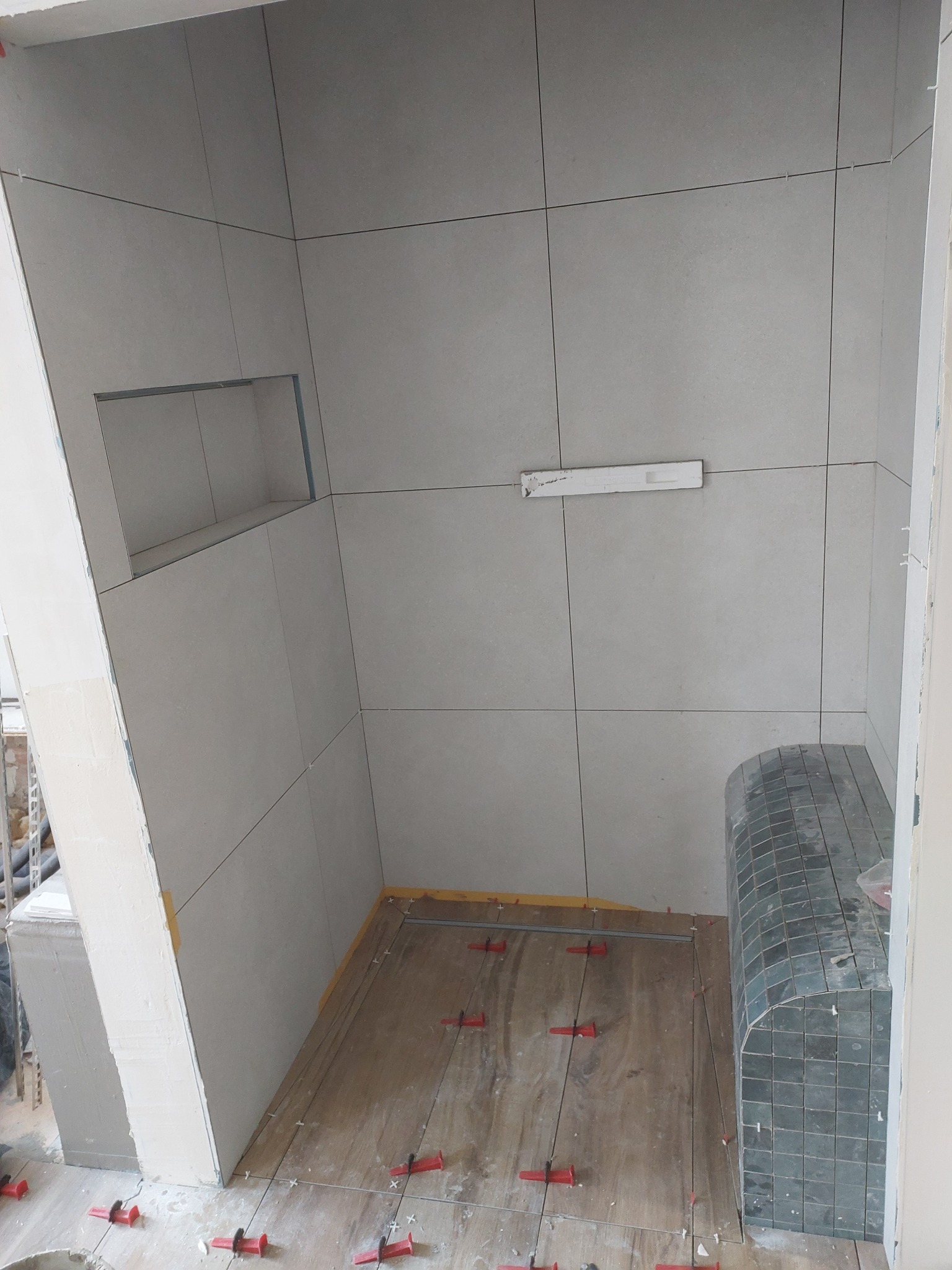 Wnętrze łazienki w trakcie prac wykończeniowych. Widoczne płytki na ścianach, podłodze i siedzisku. Użyte czerwone kliny do poziomowania płytek. Wnęka w ścianie.