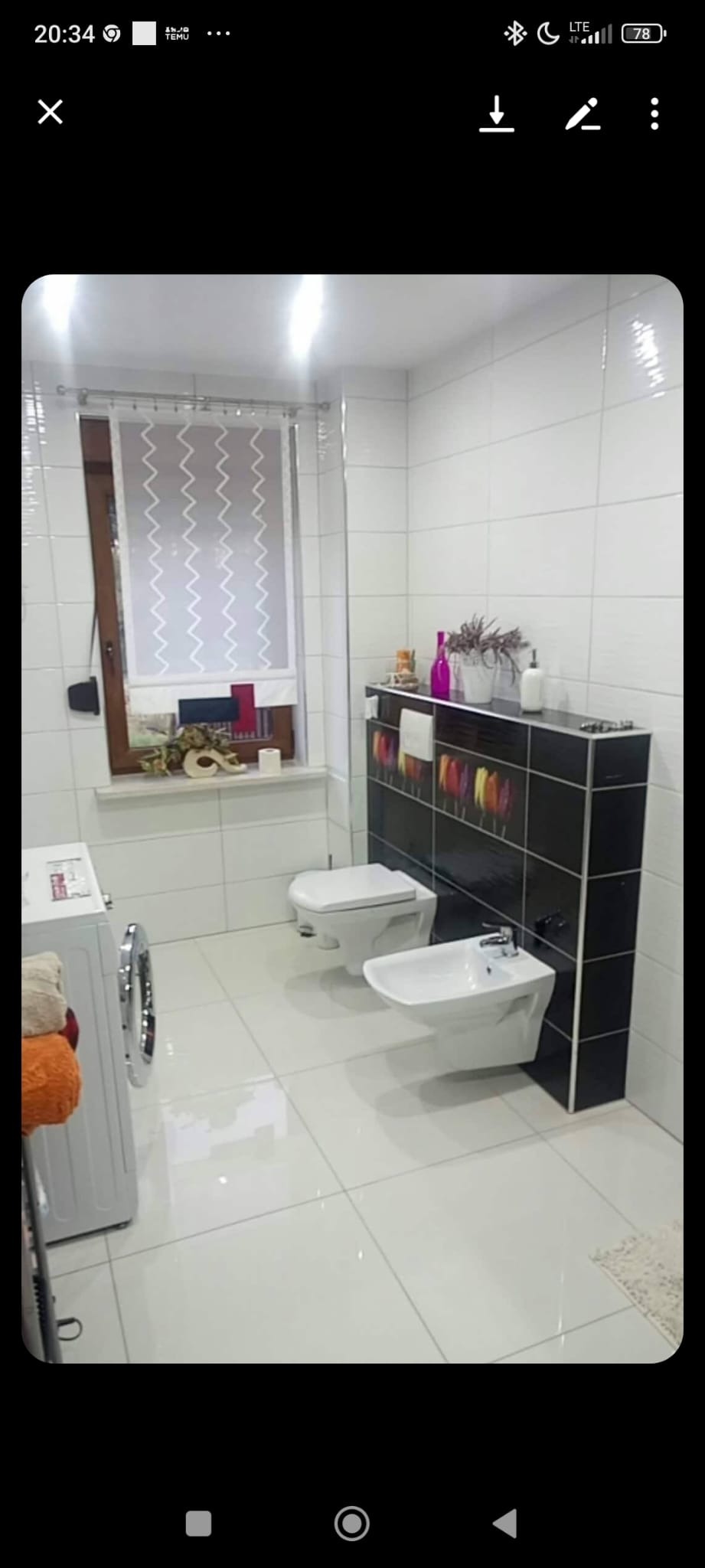 Jasna łazienka z białymi płytkami, oknem z roletą i czarną zabudową z kolorowymi akcentami. Widoczne WC, bidet i pralka. Nowoczesny design z dbałością o detale.