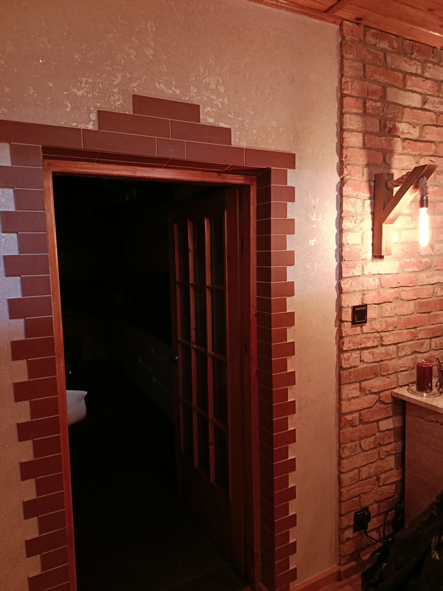 Otwarta drewniana stolarka drzwiowa z dekoracyjnym obramowaniem z cegieł, przylegająca do ściany z cegły z drewnianą lampą. Widoczny fragment wnętrza.
