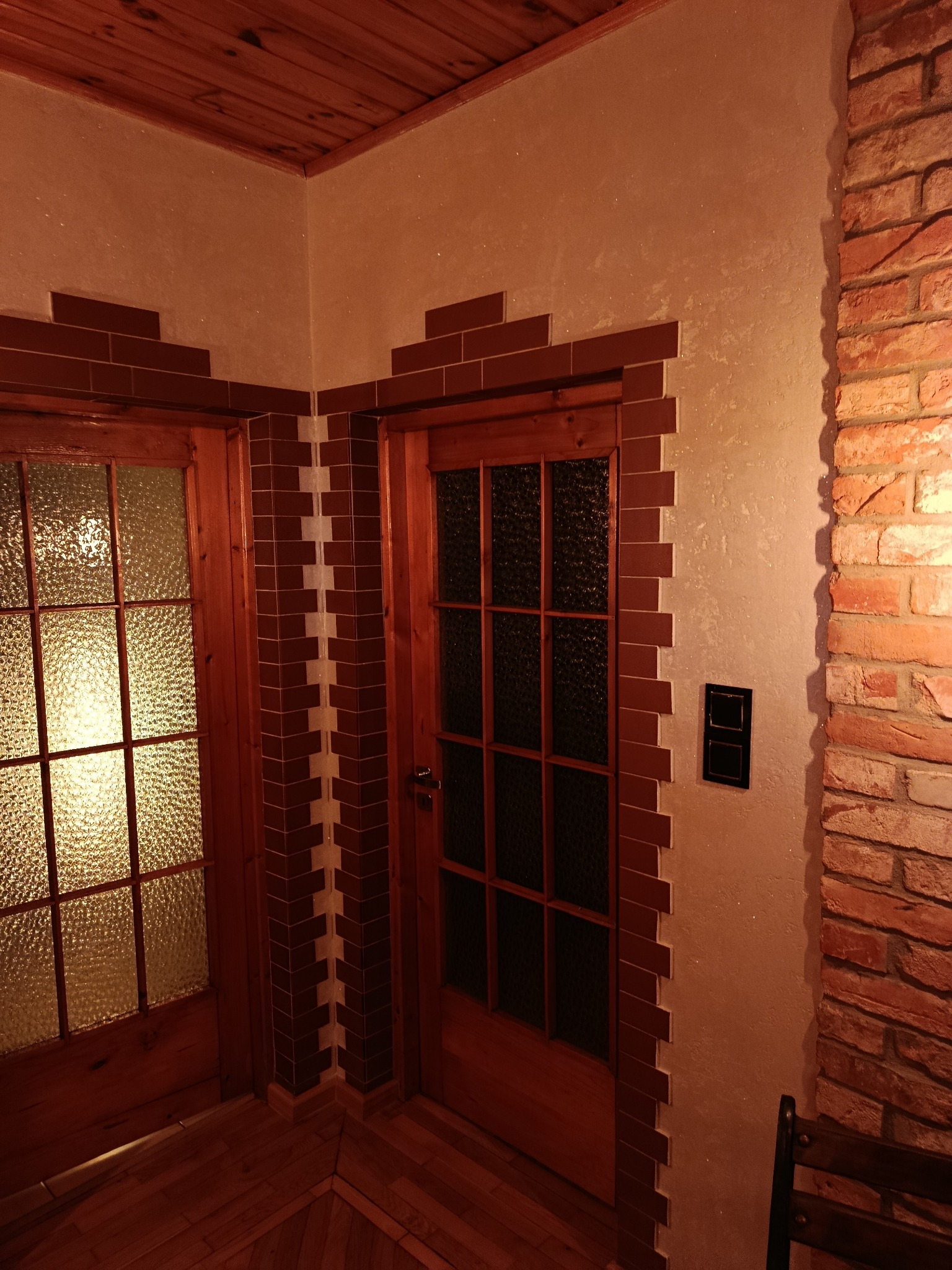 Wykończenie wnętrza z drewnianymi drzwiami, obramowanymi dekoracyjną cegłą. Ściany pokryte jasnym tynkiem strukturalnym, drewniana podłoga i sufit. Widoczny fragment ściany z cegły.
