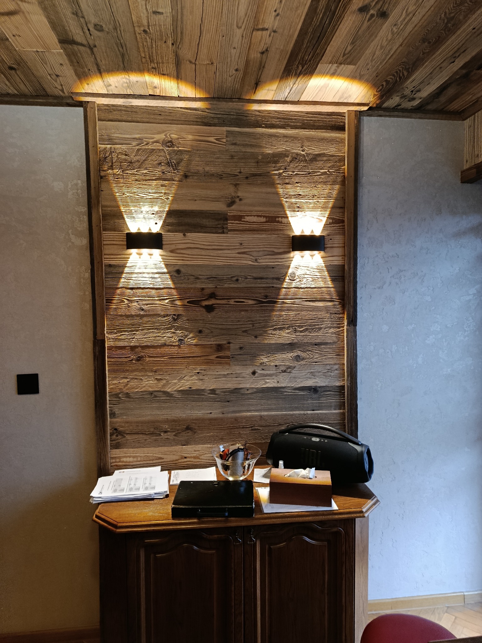 Drewniana ściana z dwoma symetrycznie umieszczonymi kinkietami, oświetlającymi przestrzeń nad komodą z akcesoriami. Ciepłe światło podkreśla fakturę drewna.