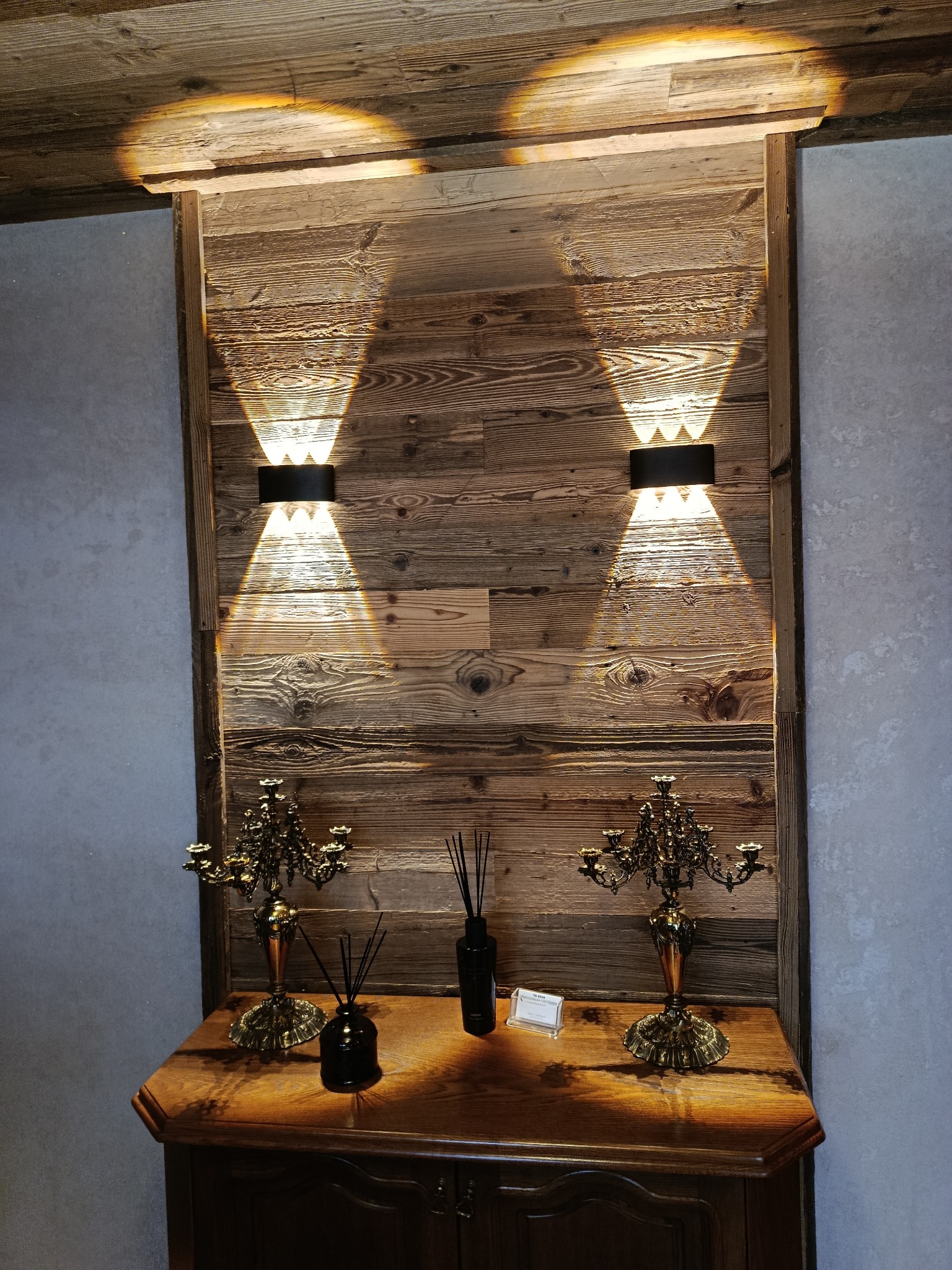 Drewniana ściana z oświetleniem punktowym i dekoracyjnymi świecznikami na komodzie. Ciepłe światło podkreśla fakturę drewna i dodaje wnętrzu przytulności.