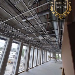 Tomasz Iwanicki GRUPA IwaTom - Sufit podwieszany segmentowy w trakcie montażu w biurze, widoczne instalacje i rusztowania. Duże okna z widokiem na zewnątrz. Logo firmy w prawym górnym rogu.