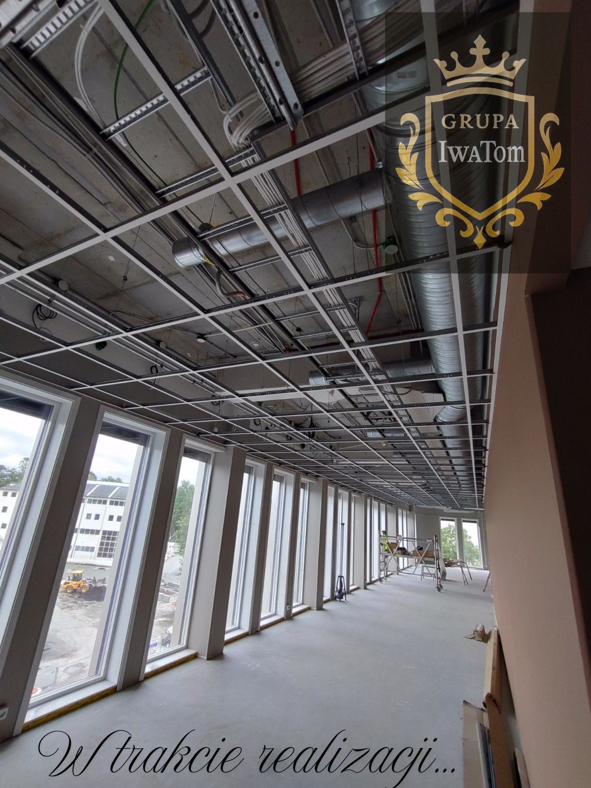 Sufit podwieszany segmentowy w trakcie montażu w biurze, widoczne instalacje i rusztowania. Duże okna z widokiem na zewnątrz. Logo firmy w prawym górnym rogu.