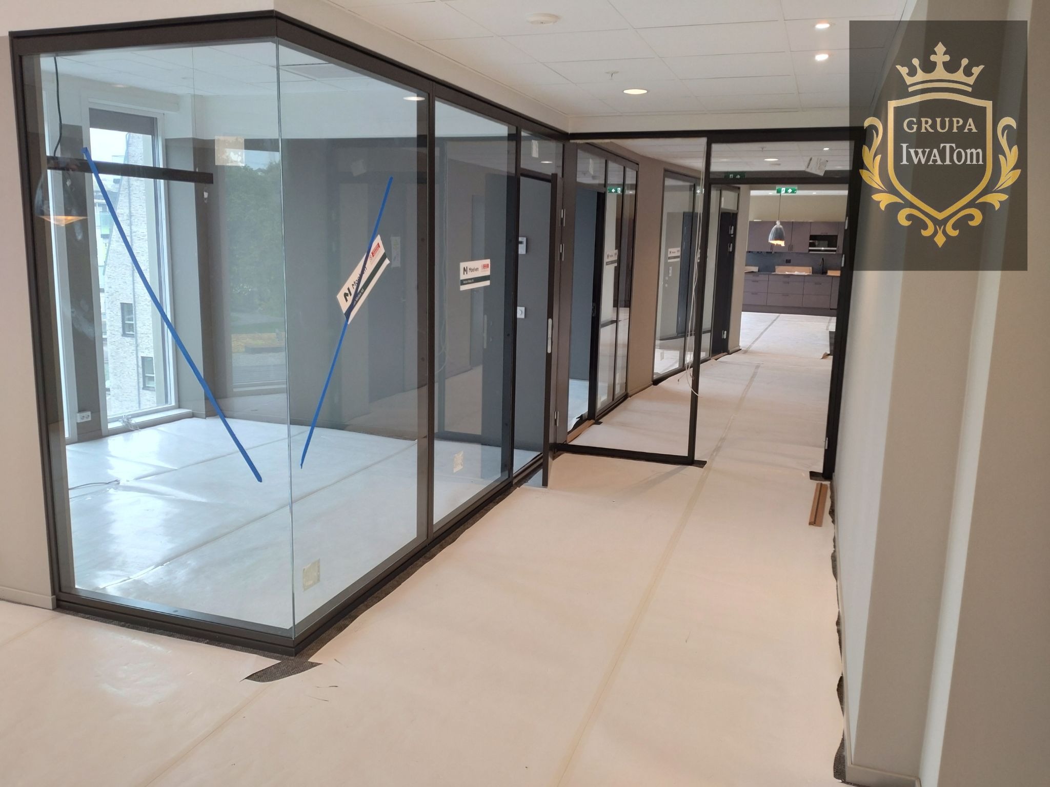 Wnętrze biura z przeszklonymi ścianami działowymi w czarnych ramach. Podłoga zabezpieczona folią ochronną, widoczne logo firmy na ścianie.