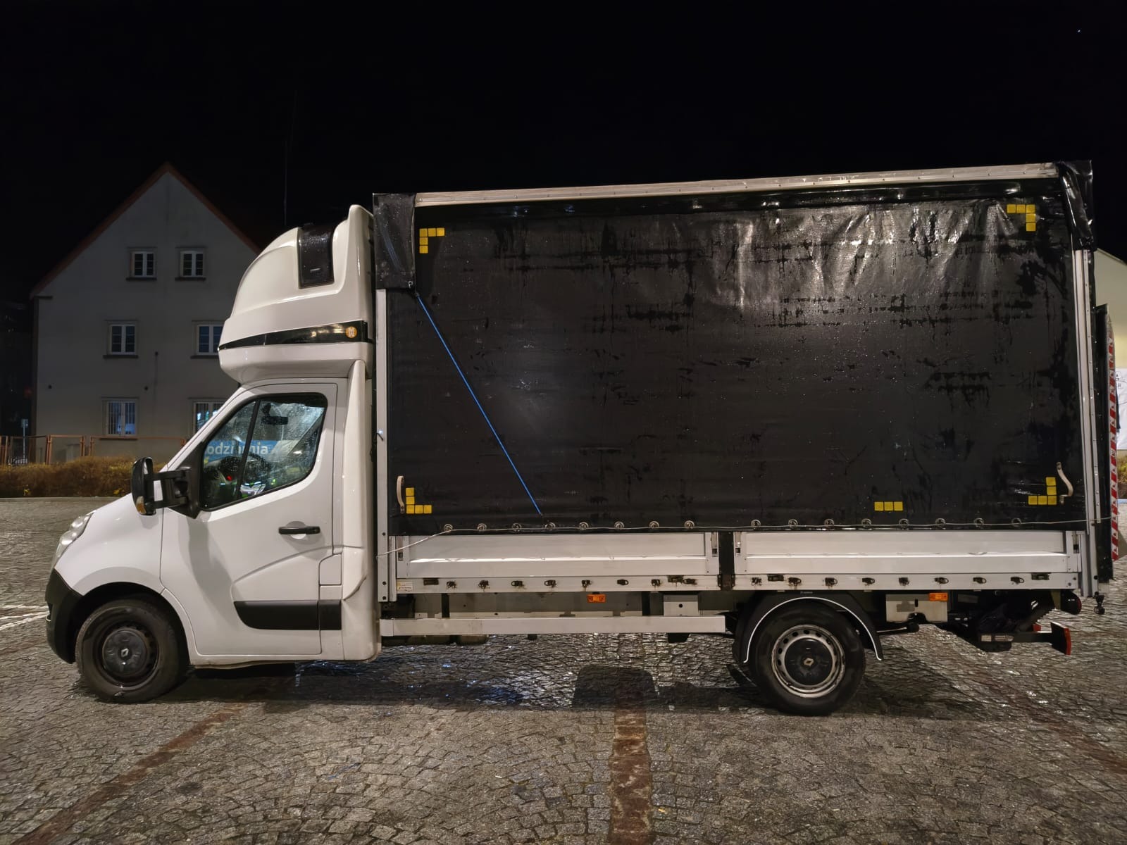 Biały samochód dostawczy z czarną plandeką, widok z boku na tle nocnego krajobrazu miejskiego. Widoczna winda załadowcza i kostka brukowa.