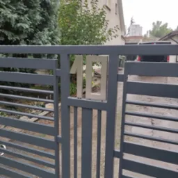 Szara brama posesji z numerem '10' wykonanym ze stali nierdzewnej, widok z bliska, w tle zieleń i fragment budynku. Lekki deszcz.