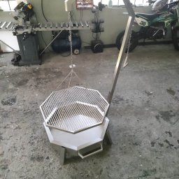 MKM WELD SERWIS - Nowoczesny grill metalowy z rusztem, zawieszony na łańcuchu, w warsztacie. Widoczny motocykl w tle i sprzęt ślusarski. Nietypowa konstrukcja grilla.