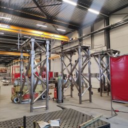 MKM WELD SERWIS - Metalowe konstrukcje schodów na hali produkcyjnej. Widoczny suwnica, stół spawalniczy z narzędziami, podnośnik nożycowy. Oświetlenie jarzeniowe.