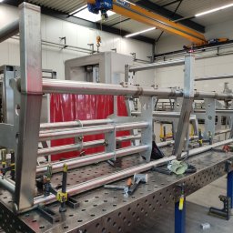 MKM WELD SERWIS - Konstrukcja metalowa spawana na stole warsztatowym, z widocznymi narzędziami i elementami mocującymi. W tle widoczna maszyna i suwnica.