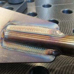 MKM WELD SERWIS - Szlifowana stal z precyzyjnymi spawami TIG, widoczne ślady termiczne i charakterystyczne tęczowe zabarwienia na spoinach. Na stole spawalniczym z otworami.