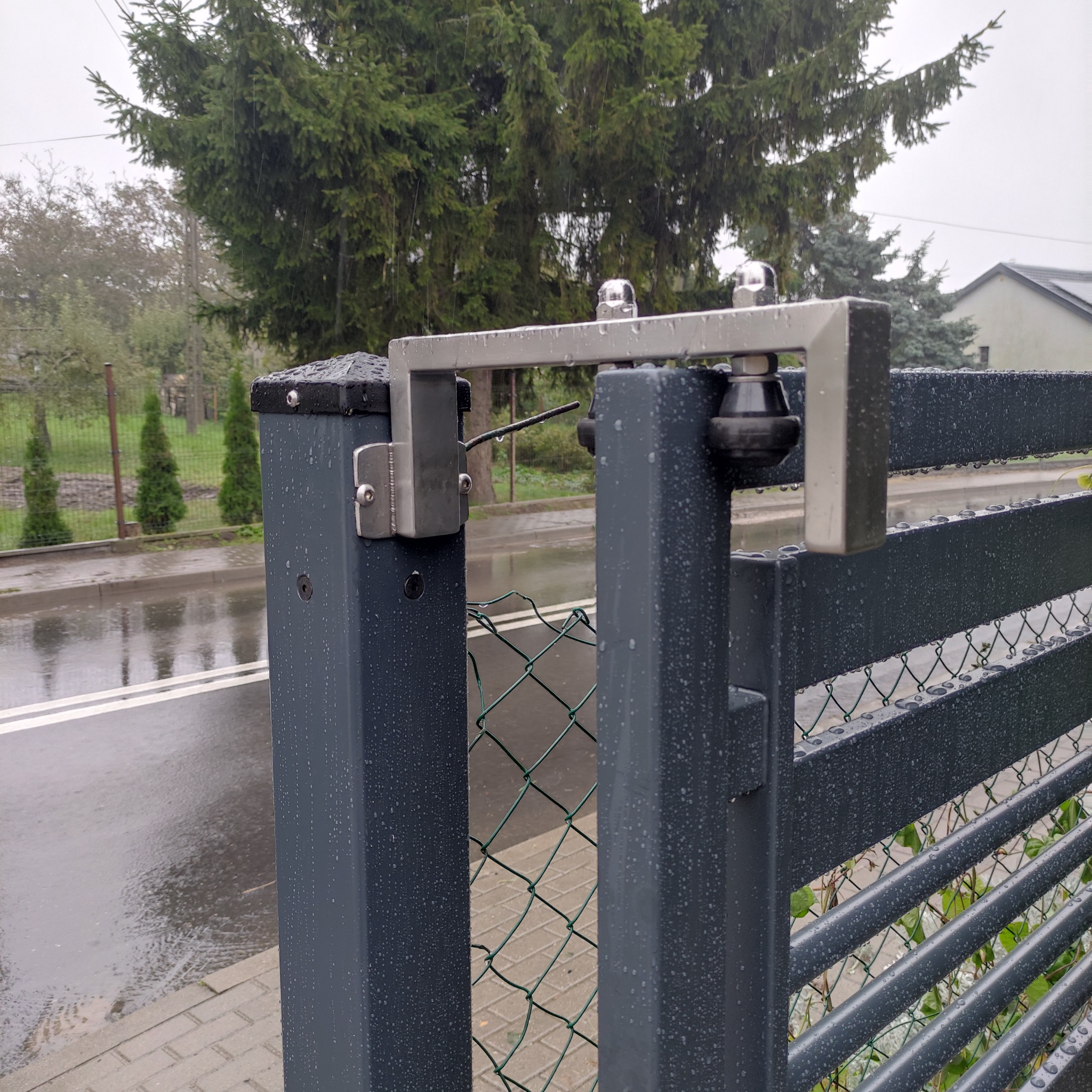 Fragment metalowej bramy wjazdowej z widocznym mechanizmem zamykającym, na tle zieleni w deszczowy dzień. Szara kolorystyka i krople deszczu na powierzchni.