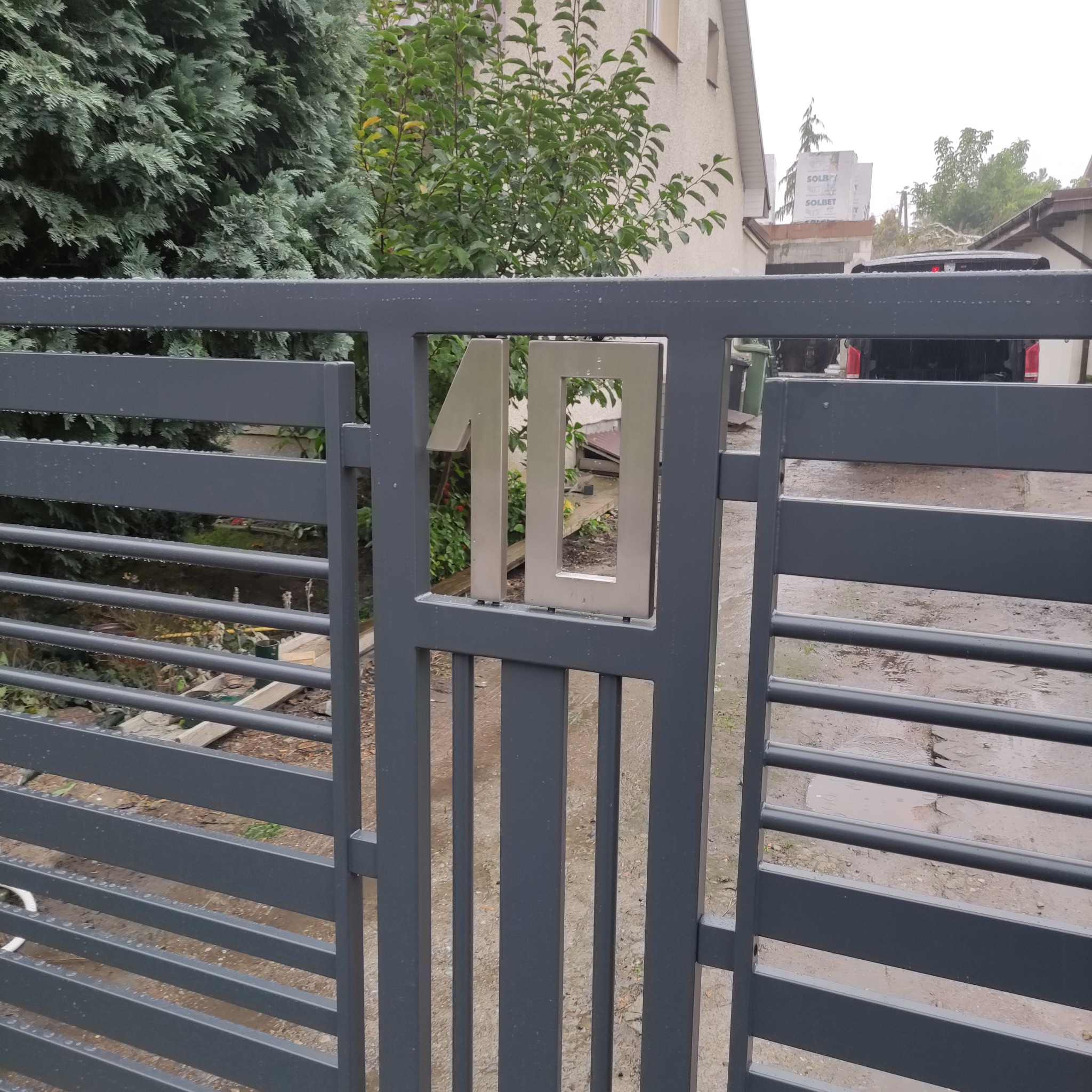 Szara brama posesji z numerem '10' wykonanym ze stali nierdzewnej, widok z bliska, w tle zieleń i fragment budynku. Lekki deszcz.