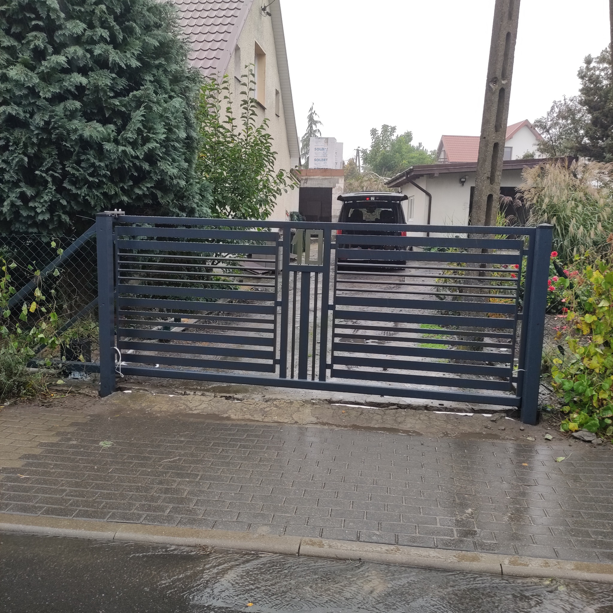 Nowoczesna, pozioma brama wjazdowa z metalowymi przęsłami, w kolorze grafitowym, na tle posesji z zielenią i budynkiem. Brama zamknięta, widoczna na mokrej nawierzchni.