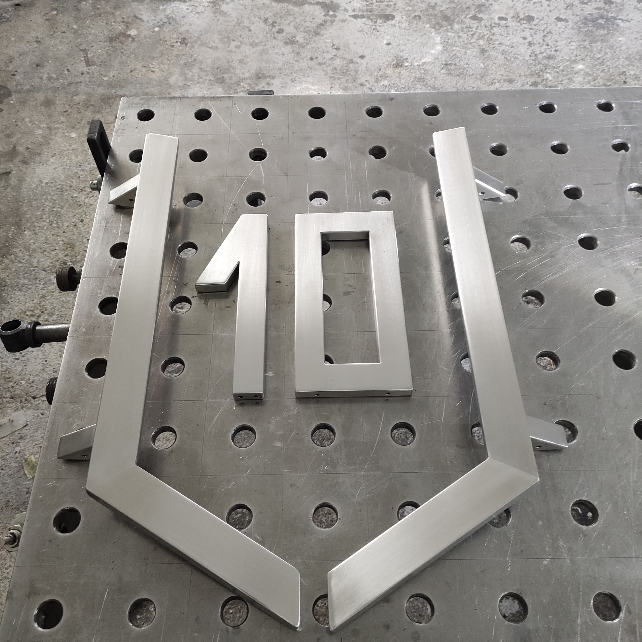 Metalowe elementy konstrukcyjne, przypominające numer '10' i fragmenty obudowy, leżą na perforowanym stole warsztatowym. Elementy są wykonane z jasnego, szczotkowanego metalu.