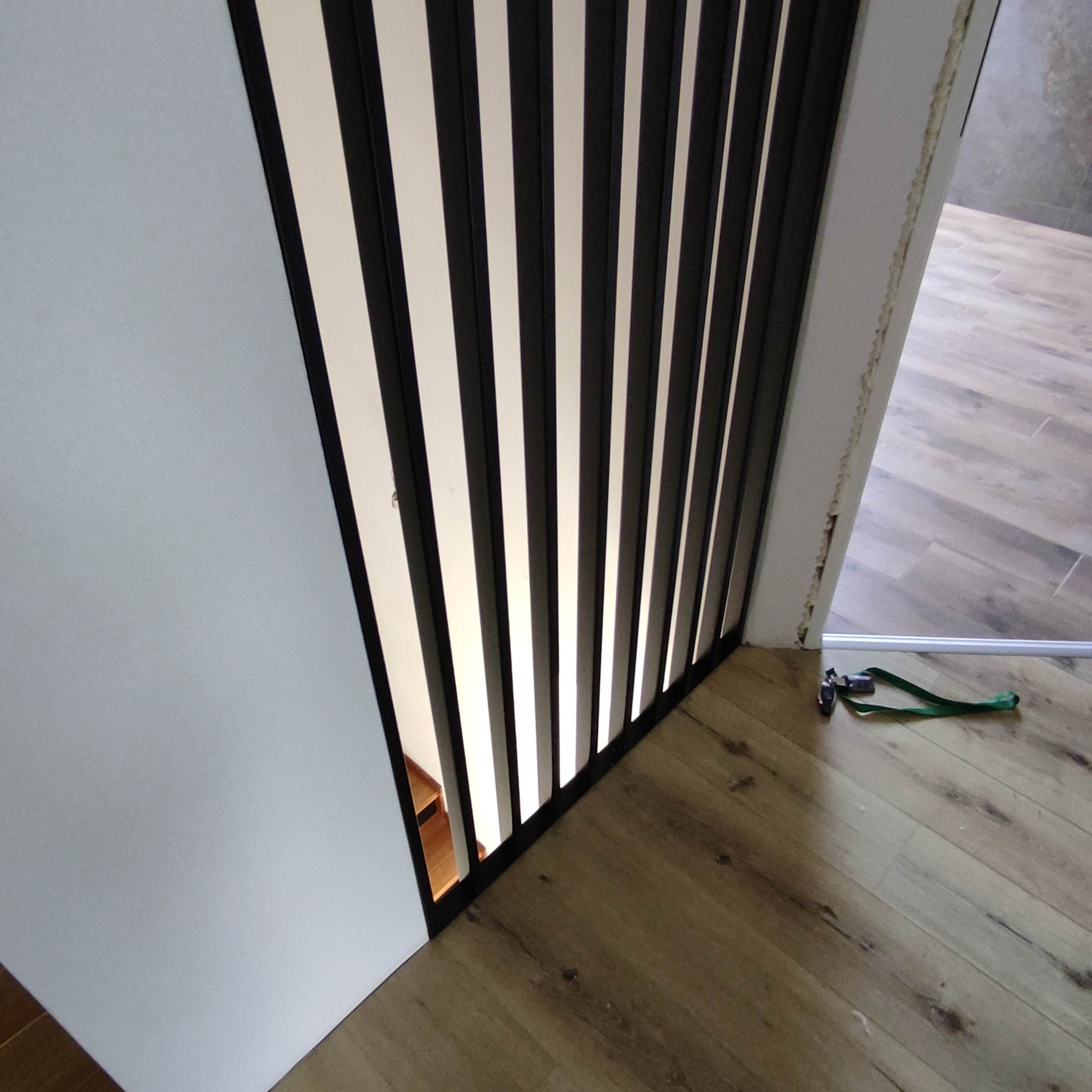 Nowoczesna balustrada schodowa z czarnymi, pionowymi szczeblami i białymi przestrzeniami, zamontowana przy ścianie. Widoczna podłoga i fragment schodów.