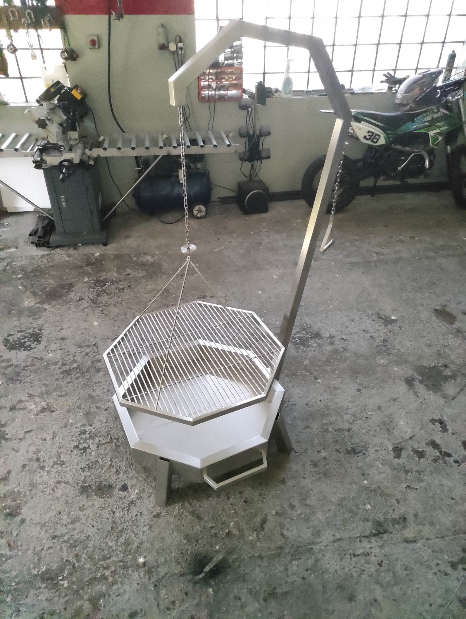 Nowoczesny grill metalowy z rusztem, zawieszony na łańcuchu, w warsztacie. Widoczny motocykl w tle i sprzęt ślusarski. Nietypowa konstrukcja grilla.