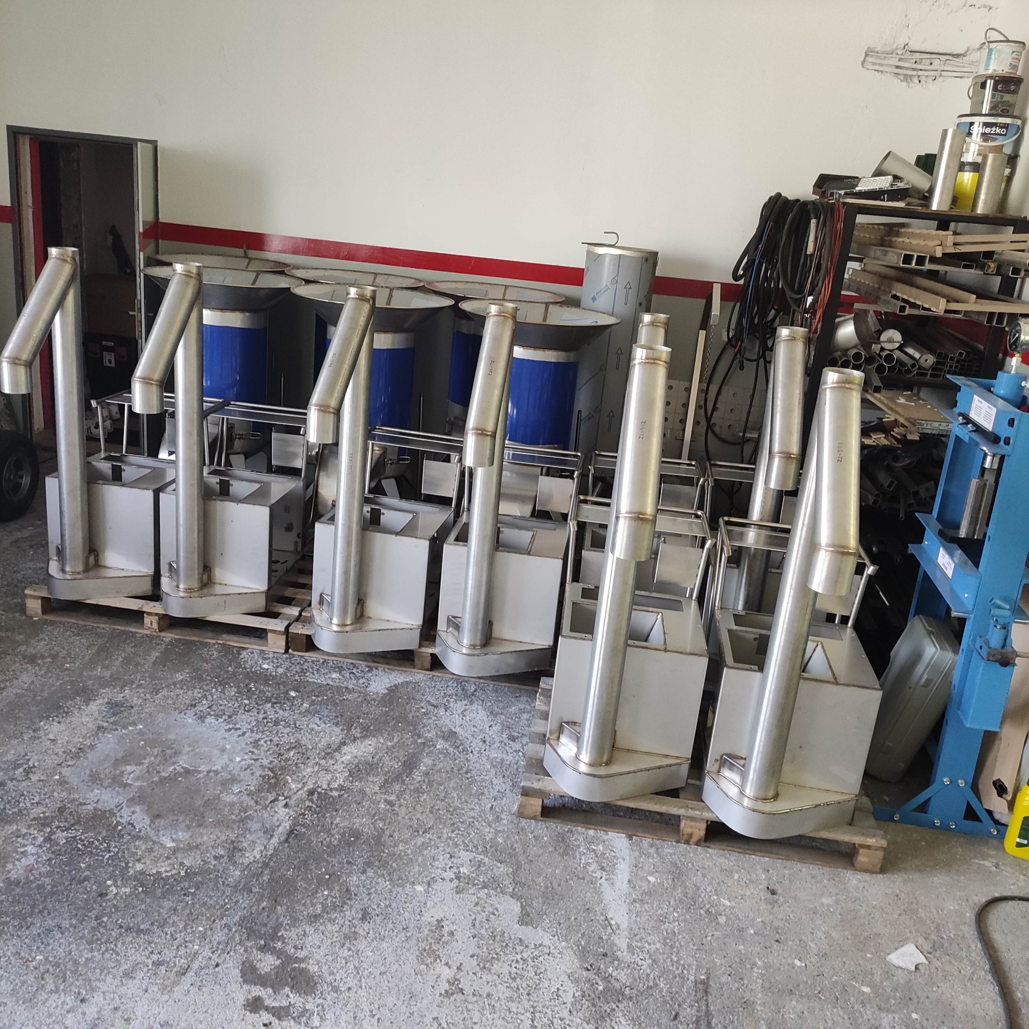 Sześć metalowych konstrukcji z rurami na paletach w warsztacie, spawane elementy, widoczne narzędzia i materiały, szare tło, produkcja przemysłowa.