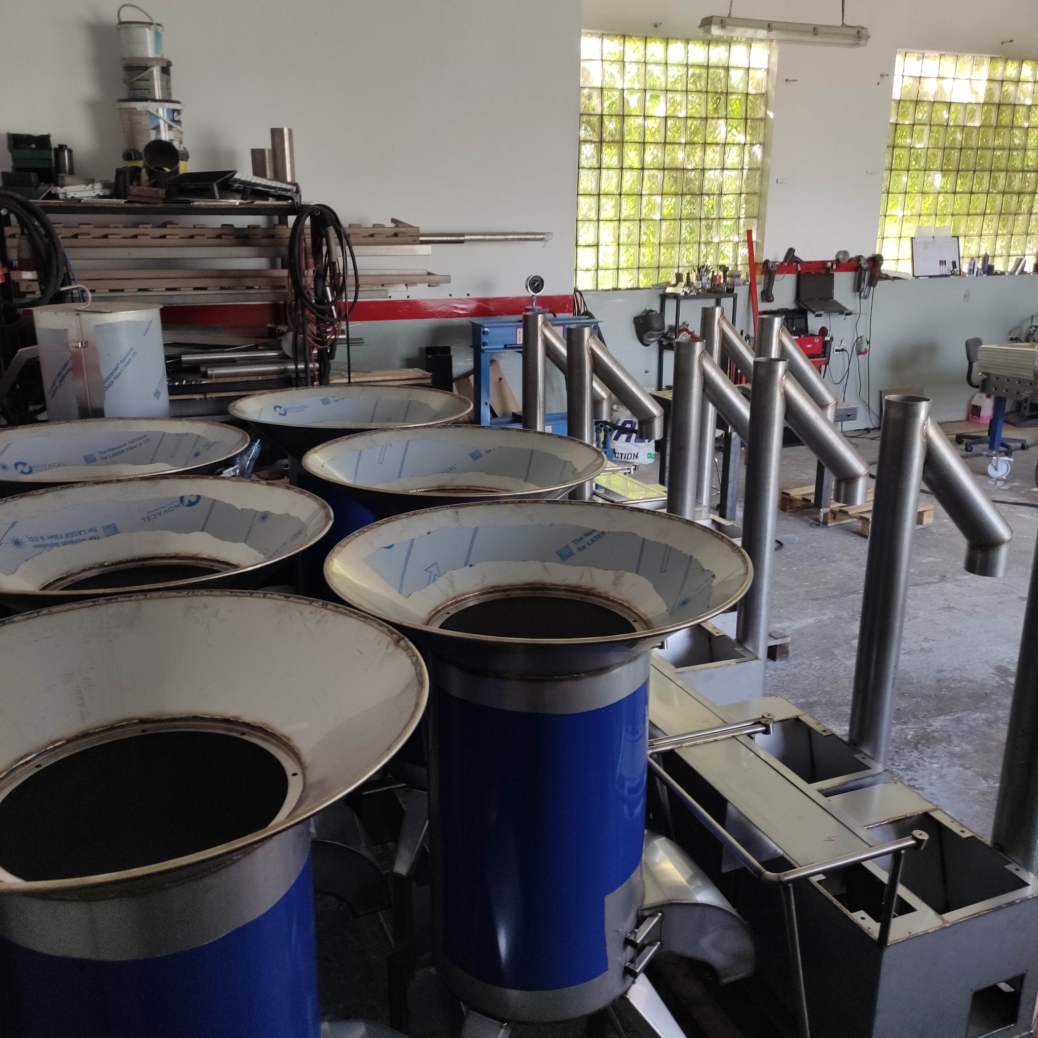 Metalowe konstrukcje w warsztacie: cylindry z kołnierzami i rury. Częściowo zdemontowane elementy z folią ochronną. Widoczne narzędzia i materiały ślusarskie w tle.