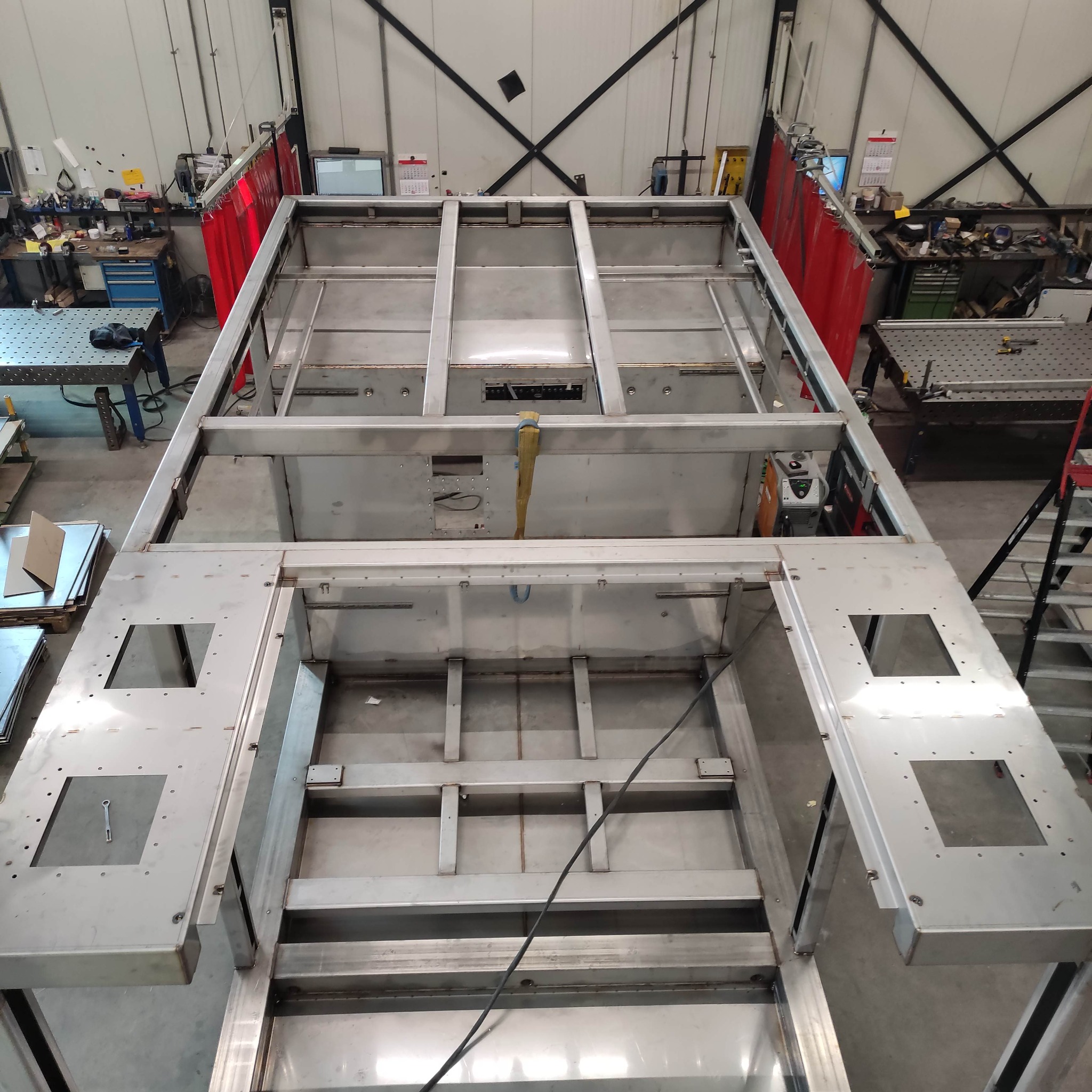 Metalowa konstrukcja w hali produkcyjnej, widok z góry. Widoczne spawane łączenia, metalowe profile i elementy mocujące. W tle stanowisko pracy spawacza z kurtyną ochronną.