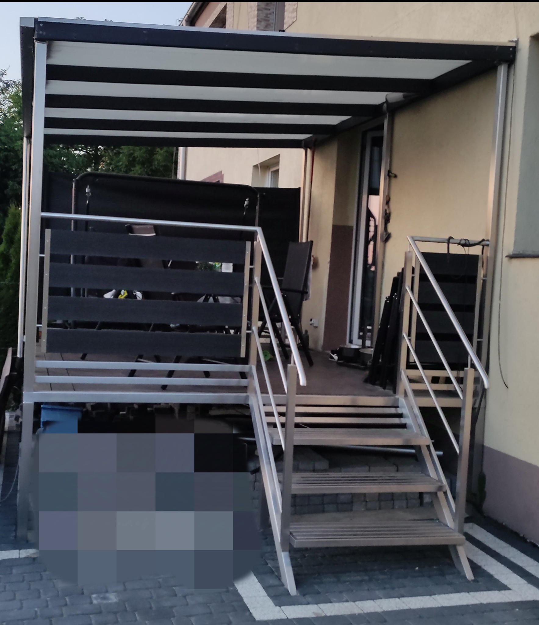 Zadaszenie balkonu z metalowymi schodami i balustradą. Konstrukcja z czarno-białym zadaszeniem, metalowe elementy w kolorze srebrnym. Widoczne elementy wyposażenia balkonu.