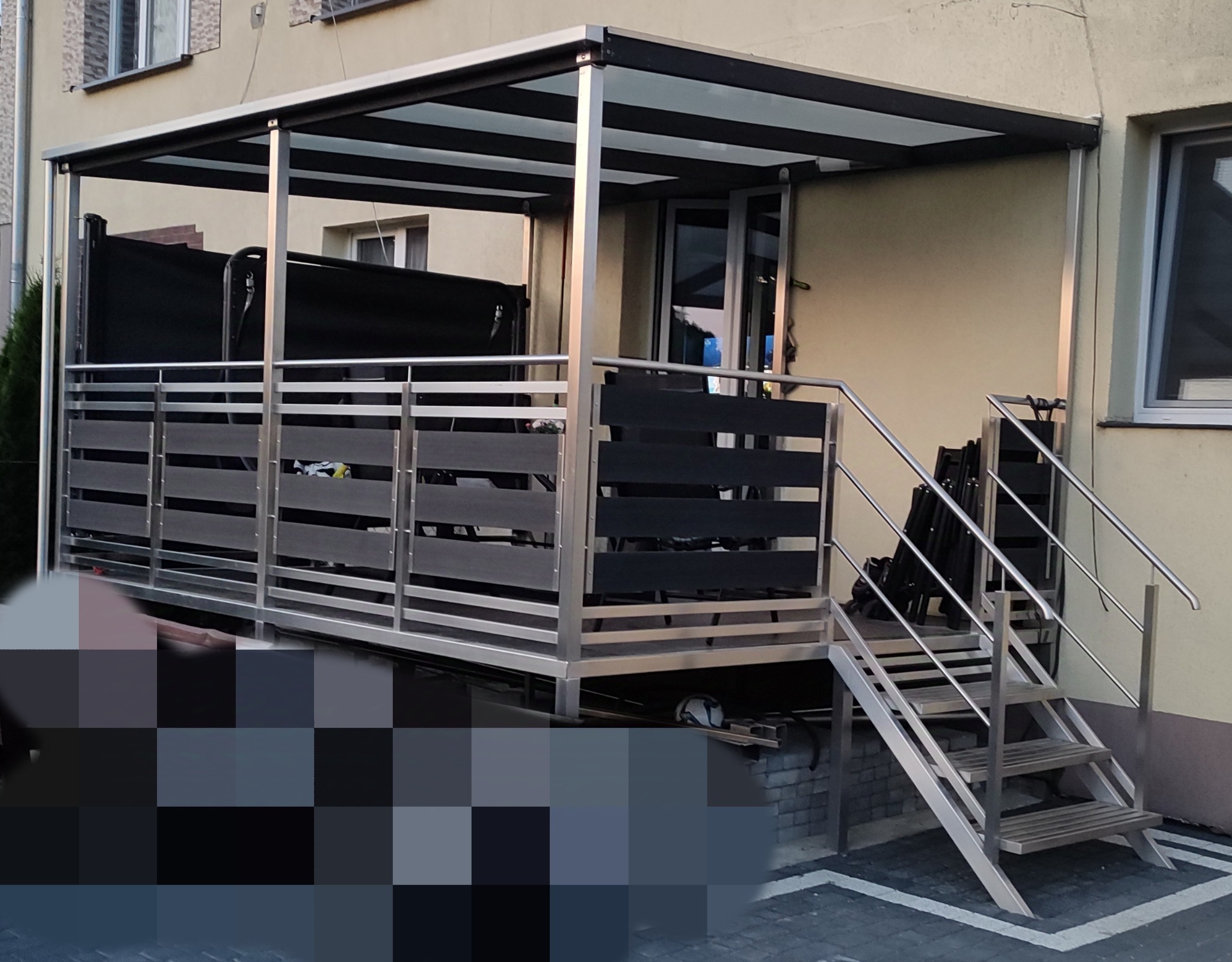 Zadaszony taras z aluminiową konstrukcją i schodami. Drewnopodobne wypełnienie balustrady. Widoczne krzesła i elementy wyposażenia. Jasna elewacja budynku w tle.