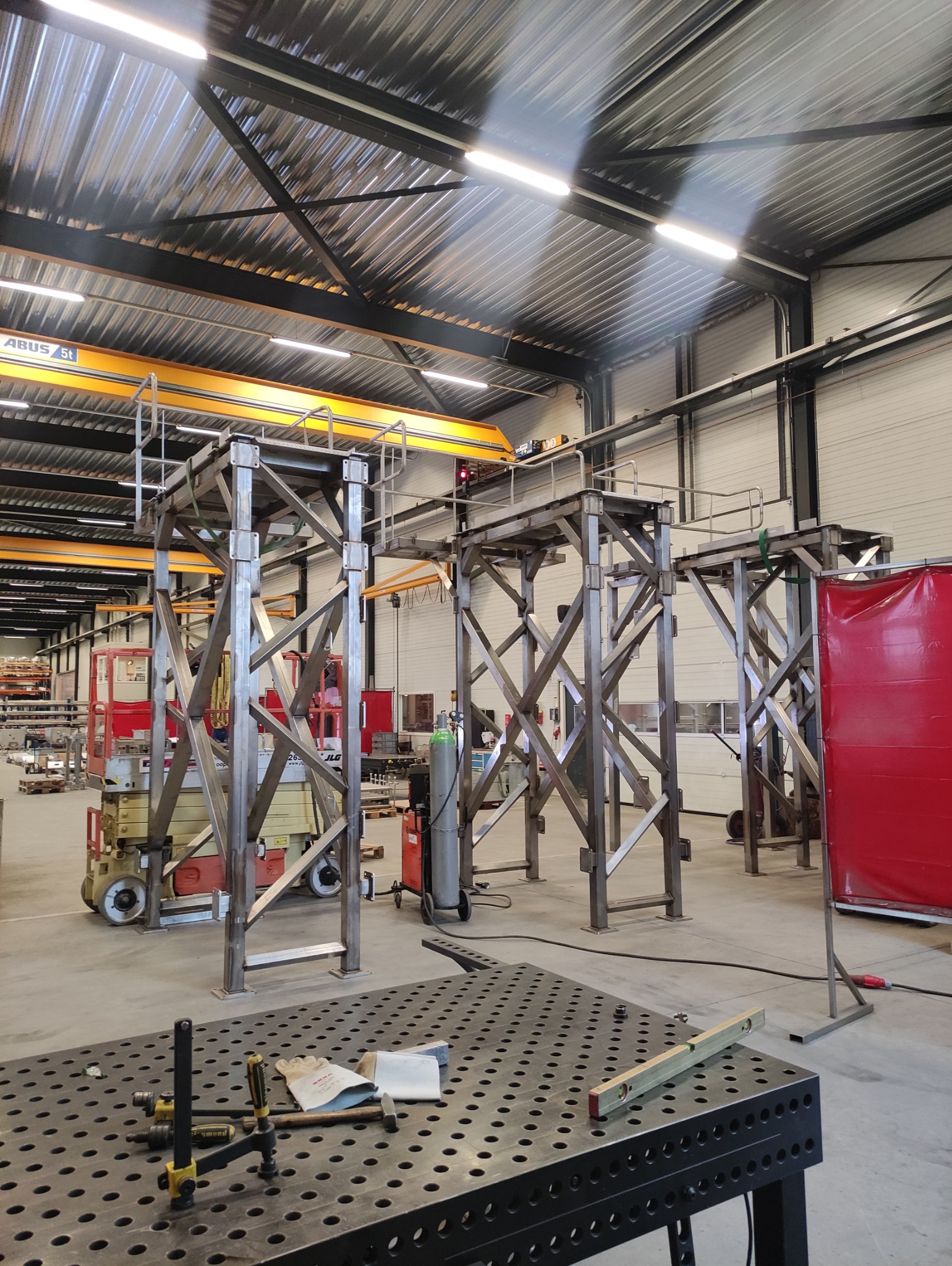 Metalowe konstrukcje schodów na hali produkcyjnej. Widoczny suwnica, stół spawalniczy z narzędziami, podnośnik nożycowy. Oświetlenie jarzeniowe.