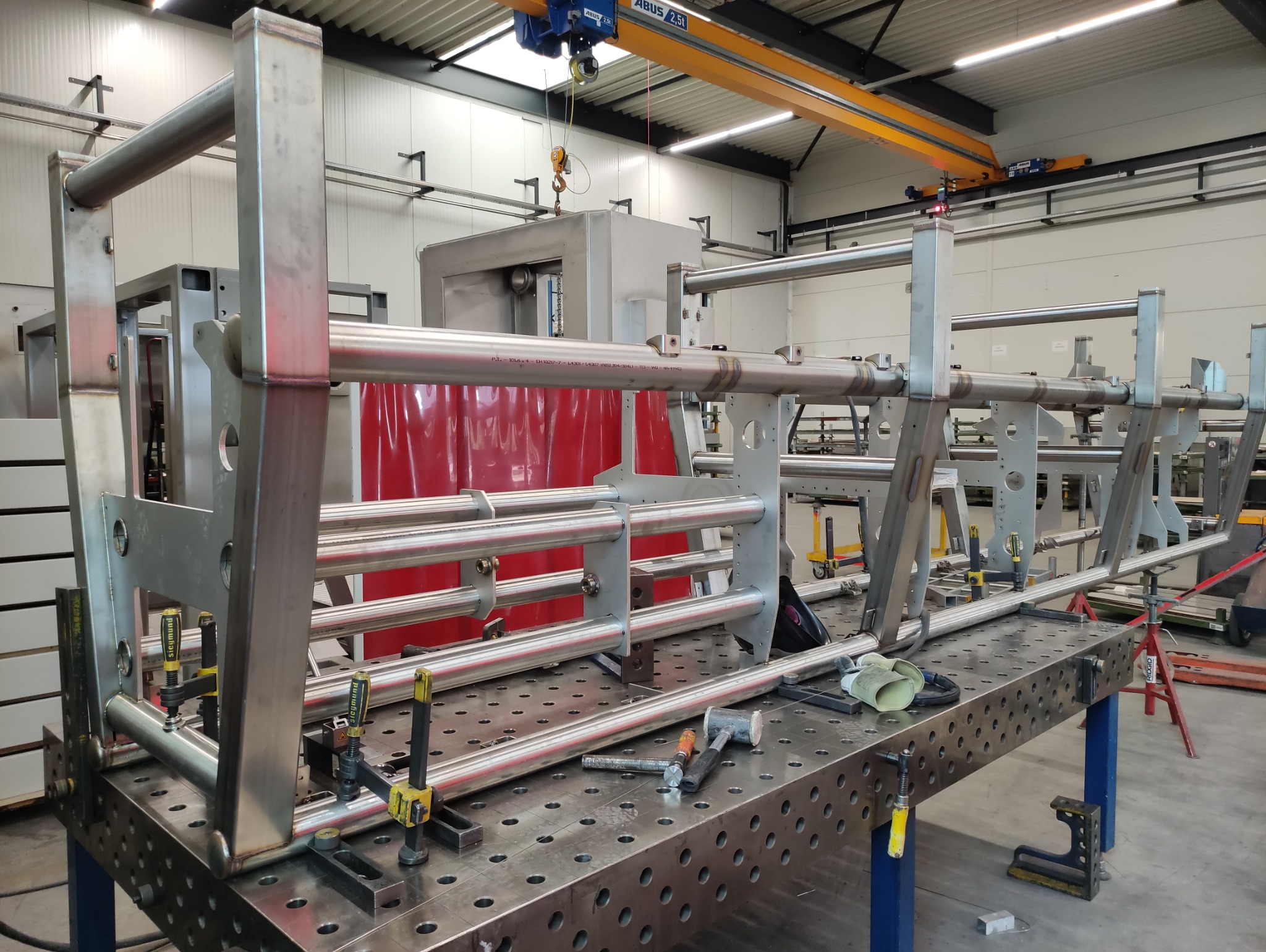 Konstrukcja metalowa spawana na stole warsztatowym, z widocznymi narzędziami i elementami mocującymi. W tle widoczna maszyna i suwnica.