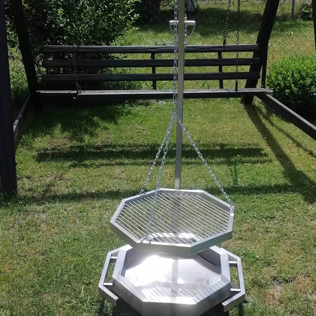 Metalowy grill ogrodowy z rusztem na łańcuchach, ośmiokątny kształt, widoczny na trawniku, w tle huśtawka.