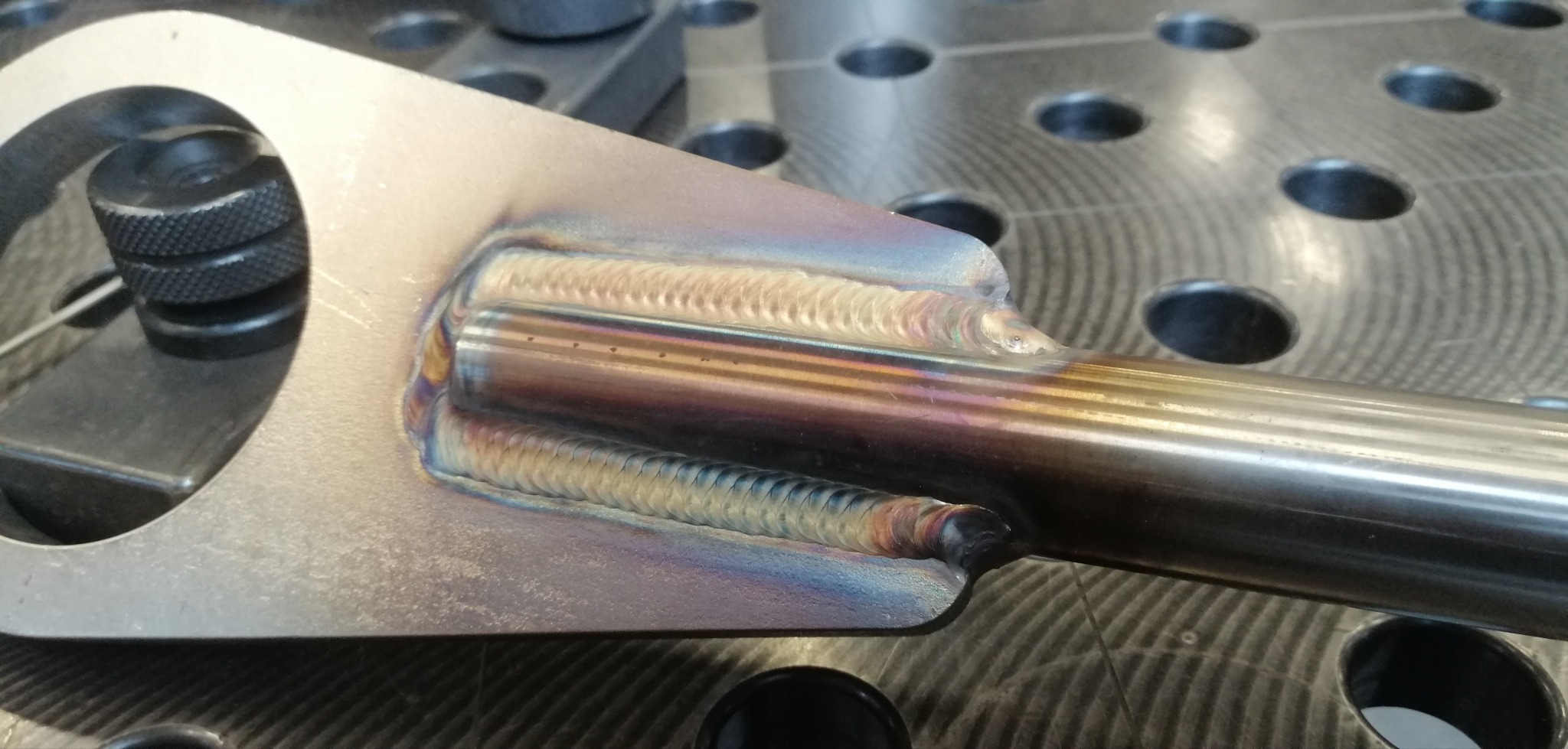 Szlifowana stal z precyzyjnymi spawami TIG, widoczne ślady termiczne i charakterystyczne tęczowe zabarwienia na spoinach. Na stole spawalniczym z otworami.