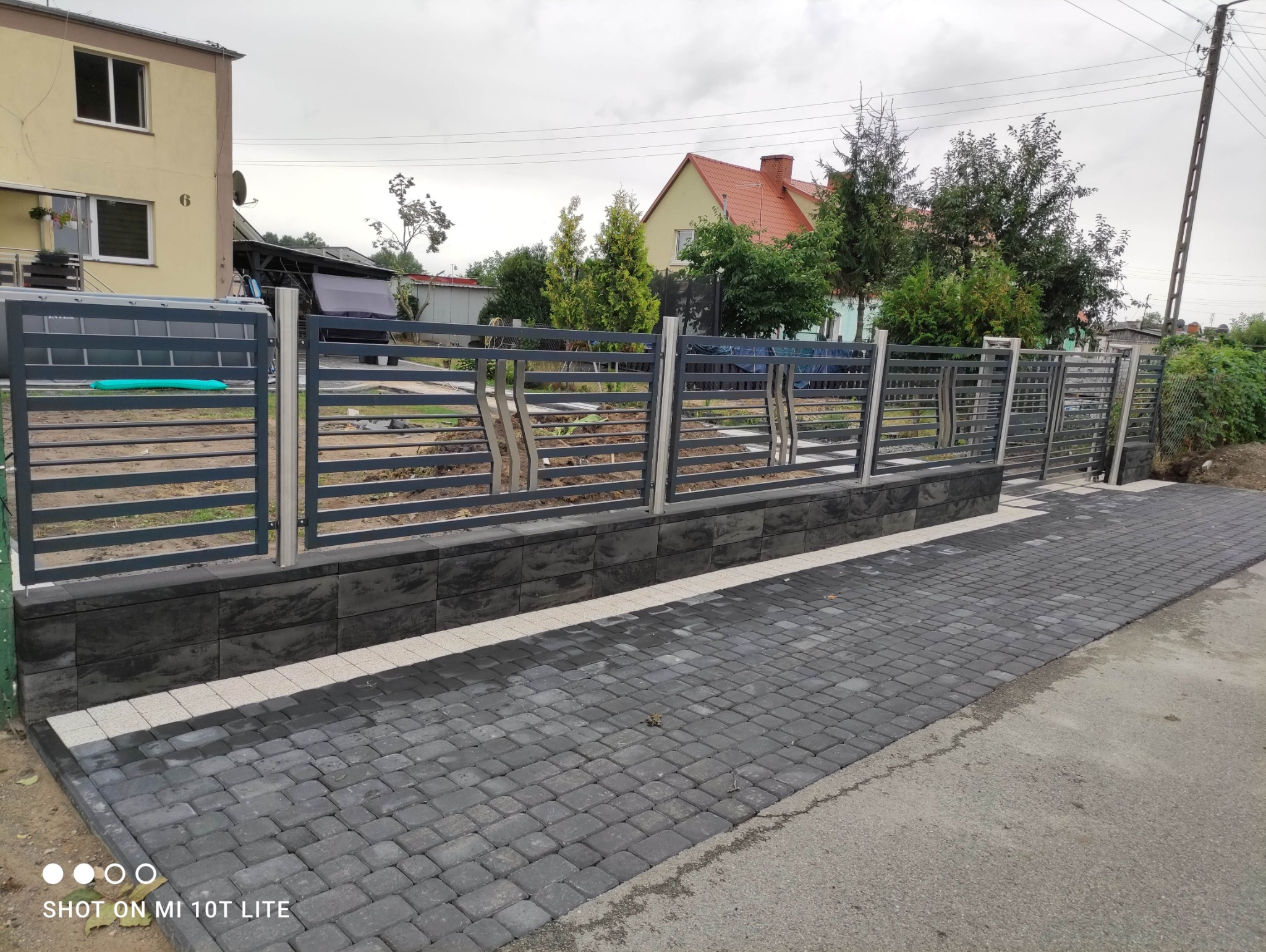 Nowoczesne ogrodzenie metalowe w kolorze grafitowym, z poziomymi przęsłami i elementami dekoracyjnymi, osadzone na niskim murku z szarej kostki. Widok z zewnątrz posesji.