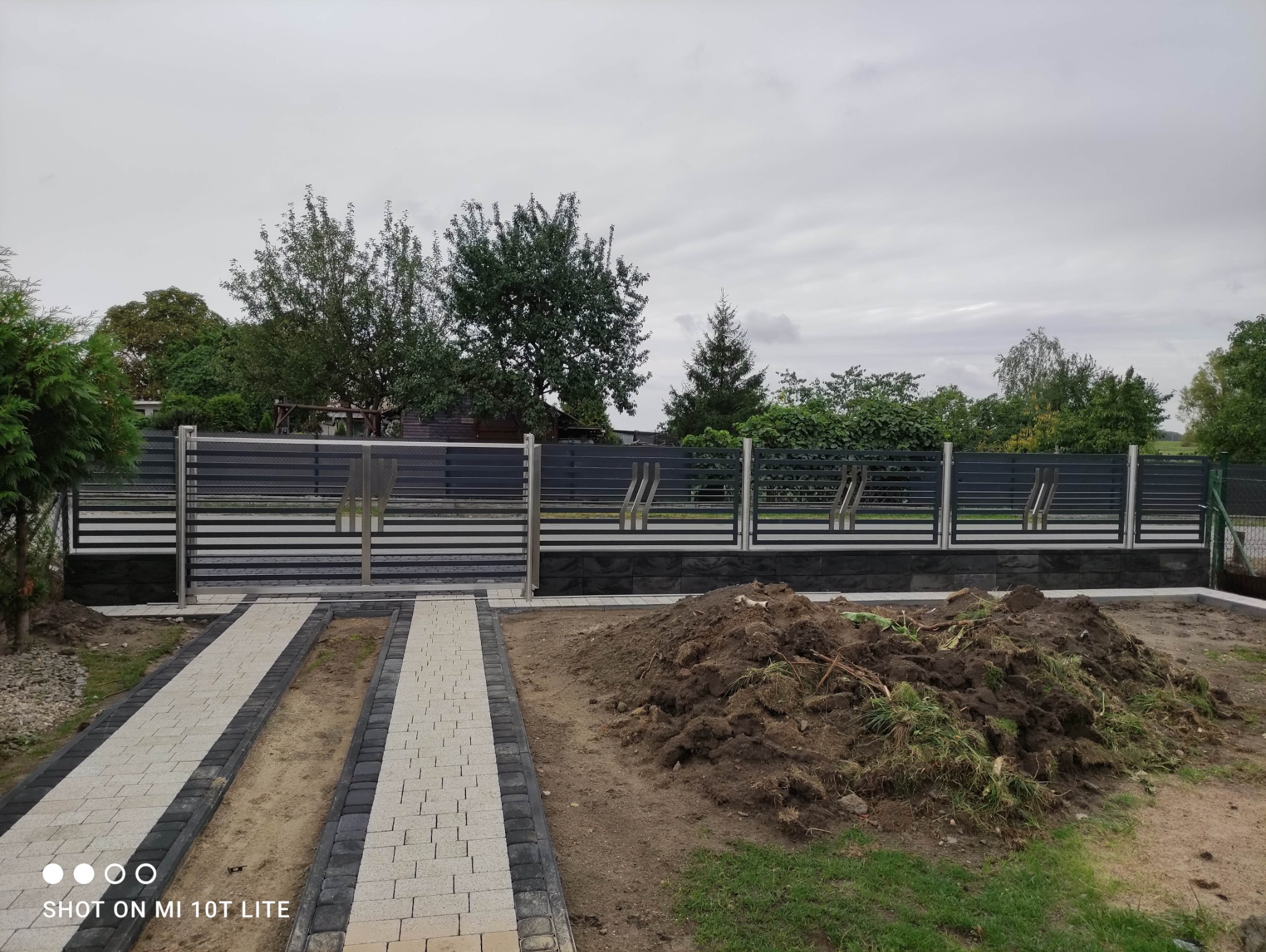 Nowoczesna brama wjazdowa z poziomymi przęsłami i stalowymi słupkami, uzupełniona o segmenty ogrodzenia. Na pierwszym planie ścieżka z kostki brukowej.