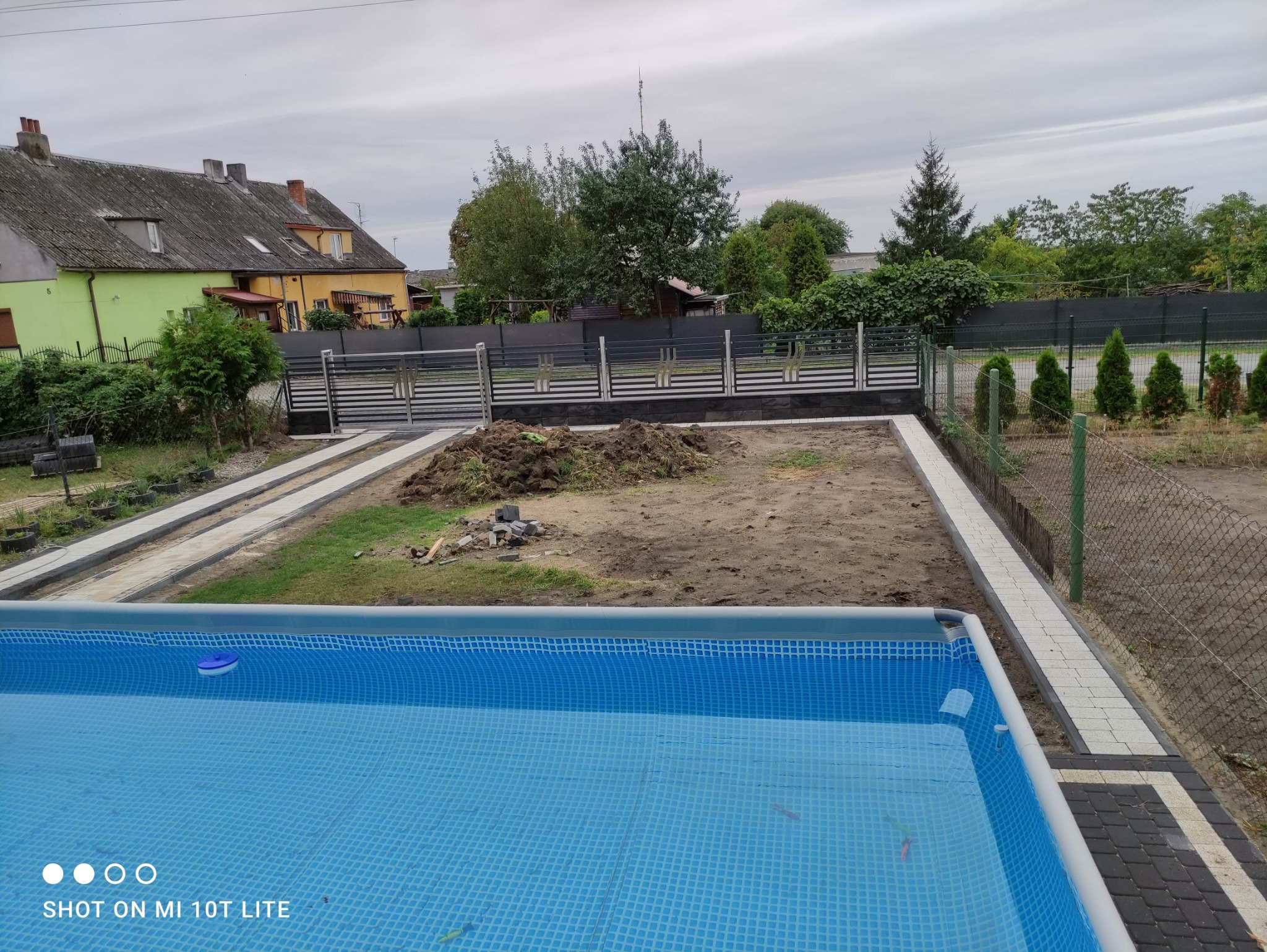 Nowoczesne ogrodzenie metalowe z elementami dekoracyjnymi, widok z perspektywy basenu ogrodowego. W tle zabudowa domów i zielone drzewa.