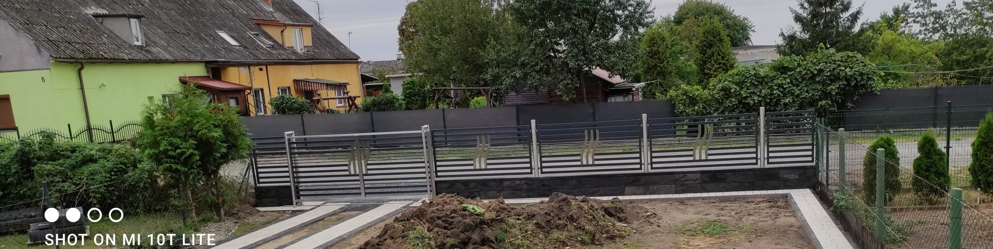 Nowoczesne ogrodzenie posesji z metalowymi przęsłami i podmurówką z kamienia łupanego. Widok na dom i ogród w tle. Elementy posesji w trakcie prac ziemnych.