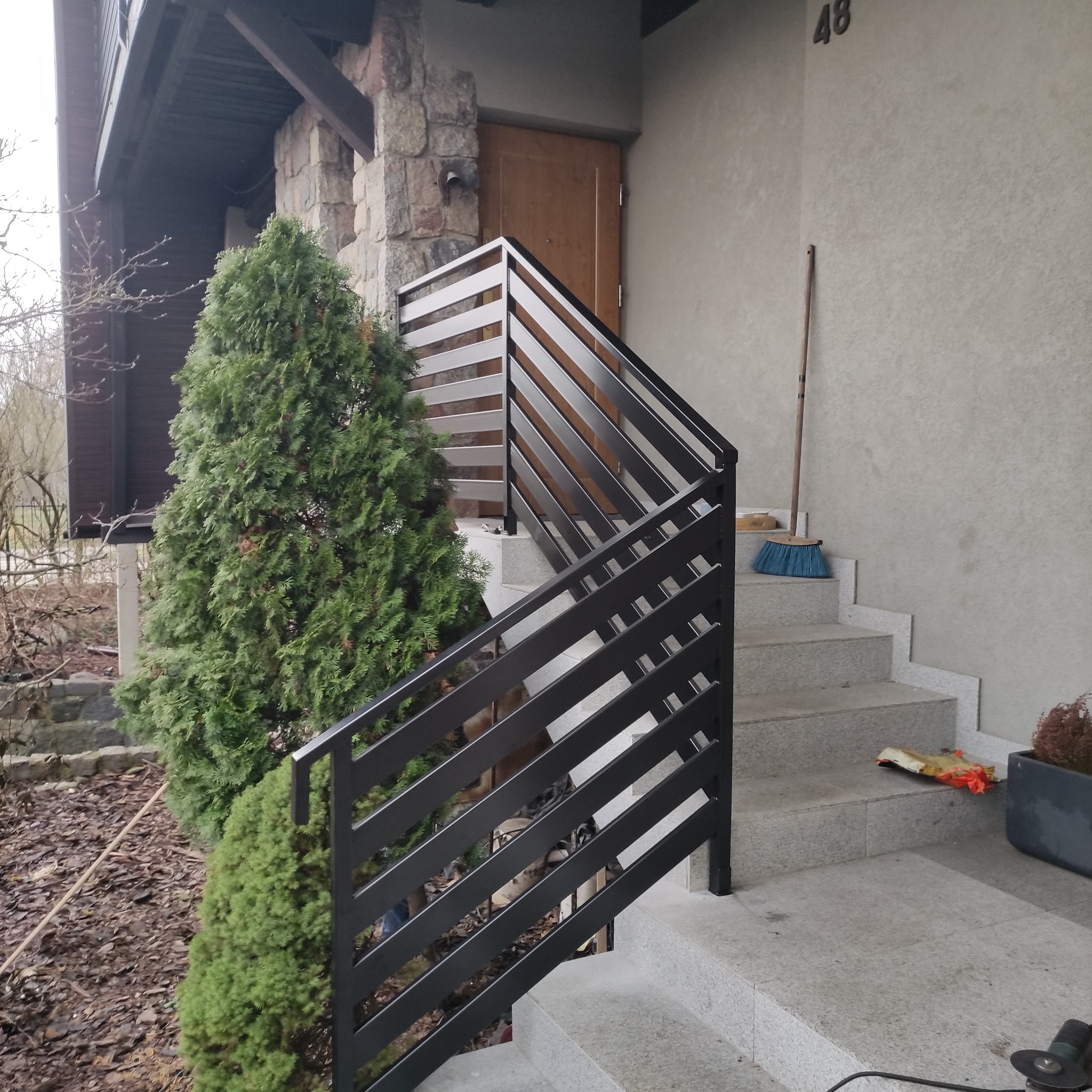 Nowoczesna, czarna balustrada schodowa z poziomymi szczeblami, zamontowana przy wejściu do domu z numerem 48. Obok rośnie zielony krzew.